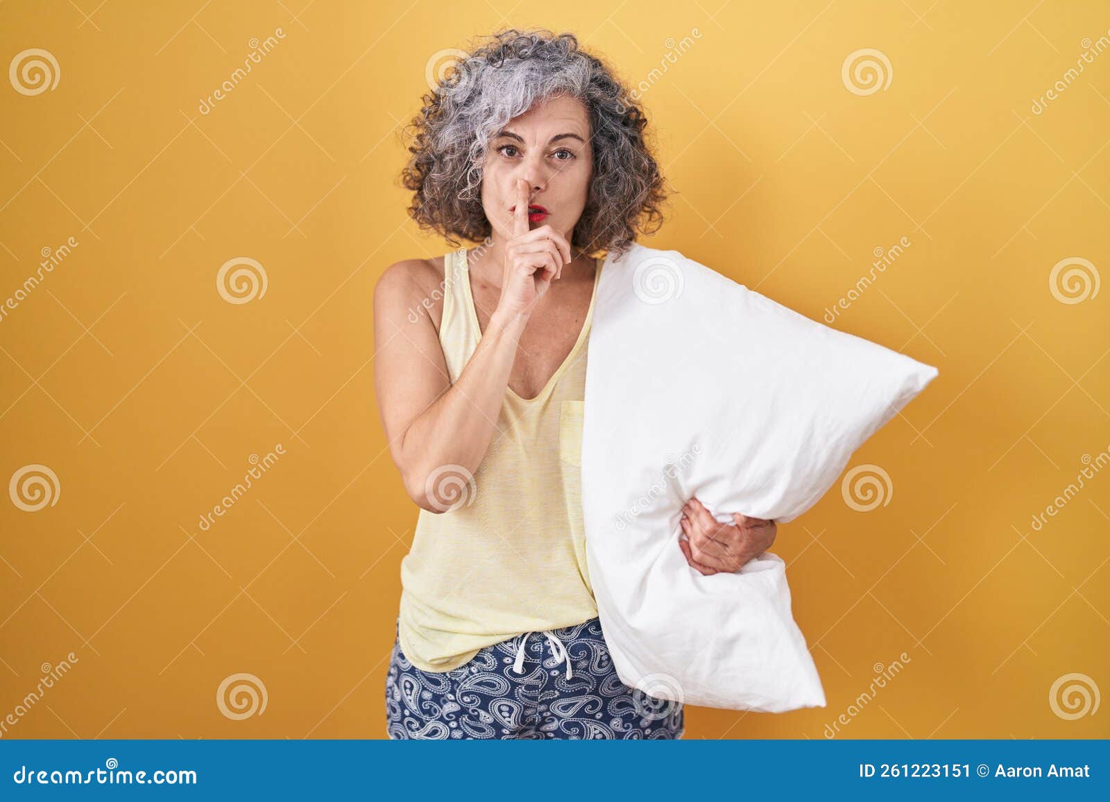 middle age woman with grey hair wearing pijama hugging pillow asking to be quiet with finger on lips