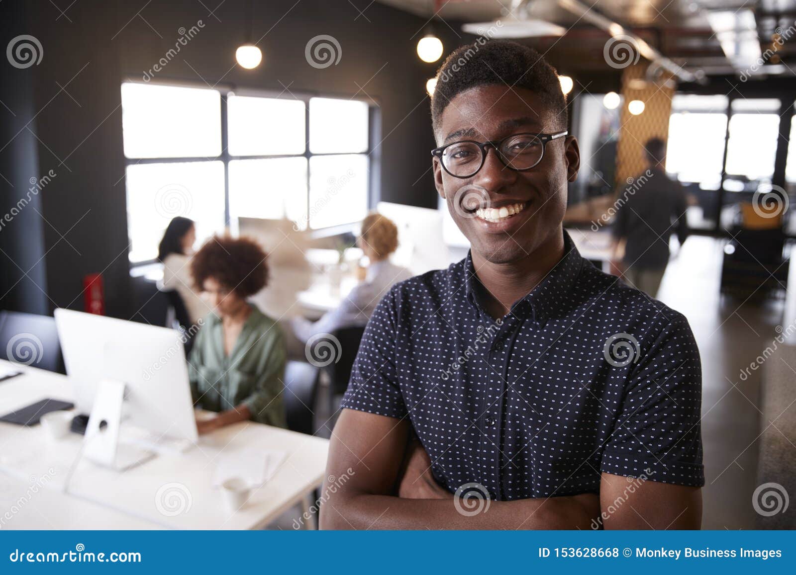 millennial black male creative standing in a busy casual office, smiling to camera