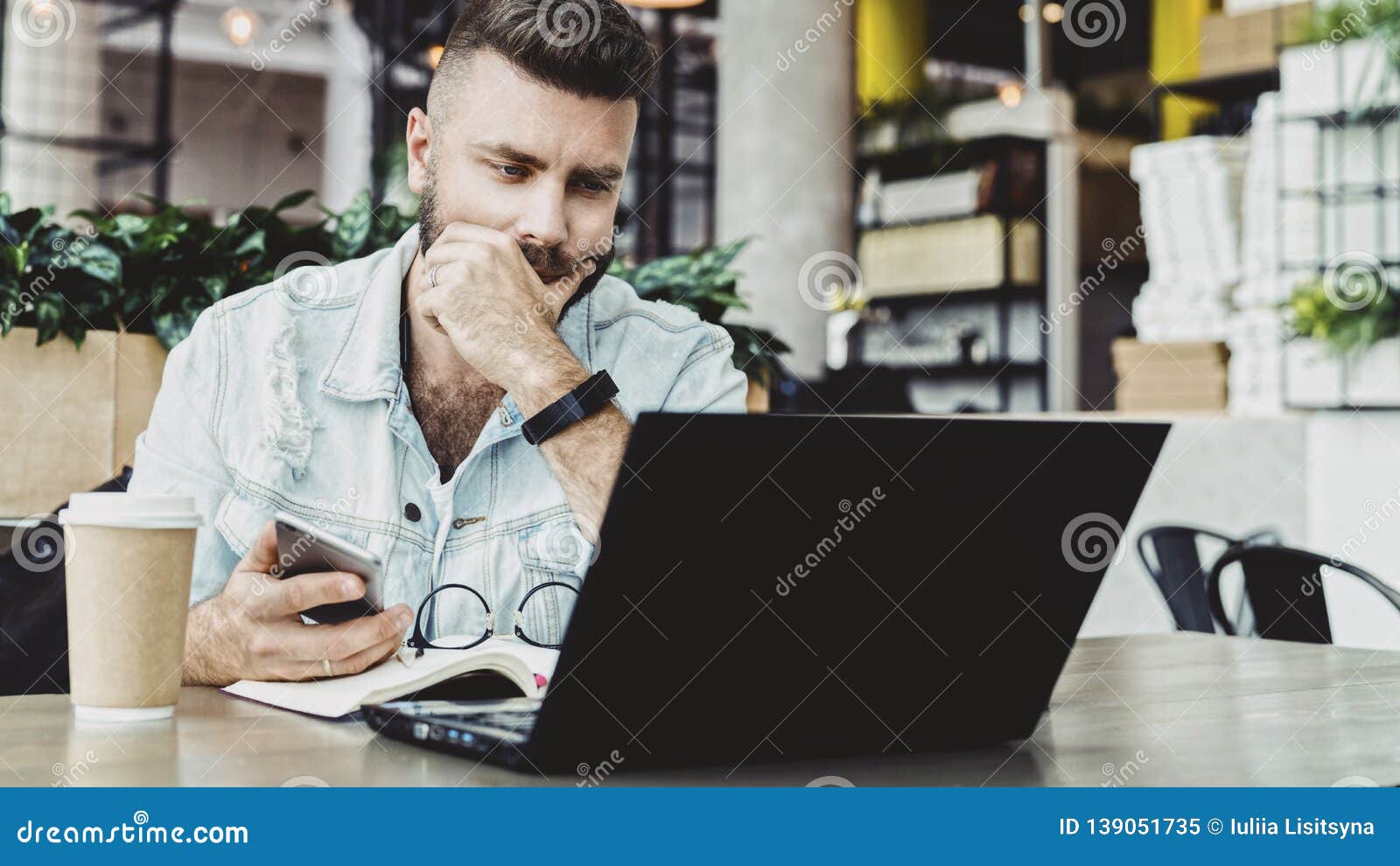 millennial businessman sitting in cafe with open laptop, looking thoughtfully at computer screen, holding smartphone.