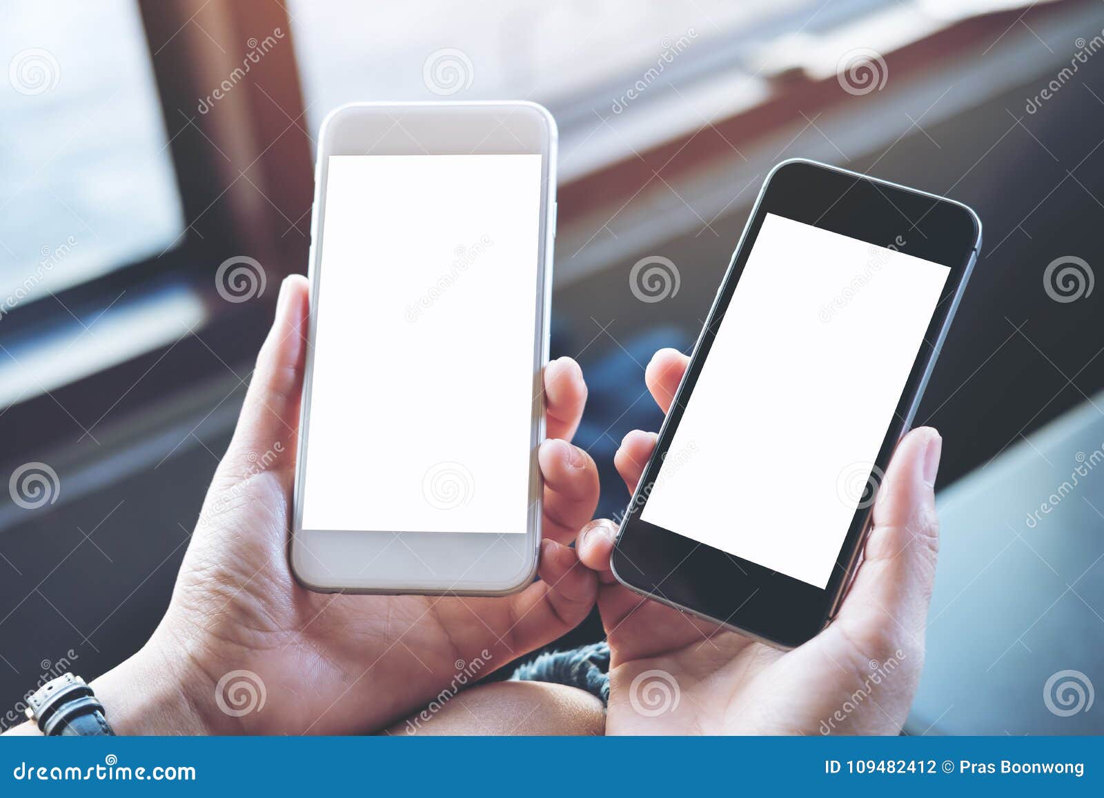 a woman`s hands holding two mobile phones with blank white screen in modern cafe