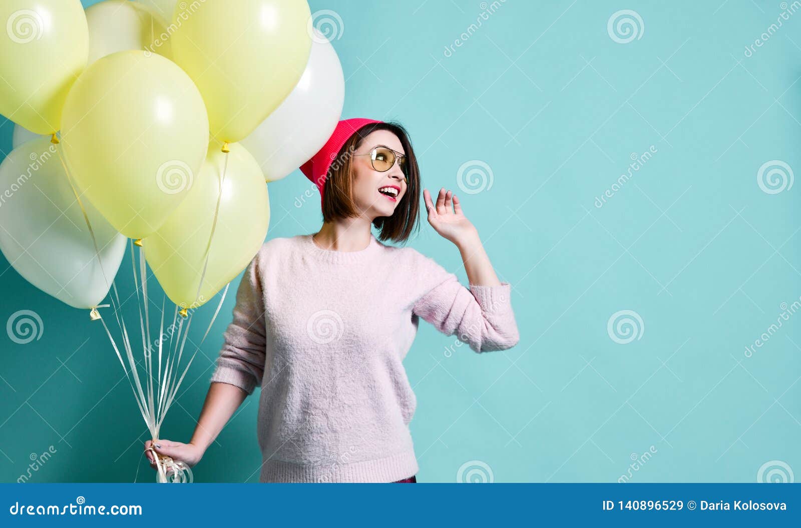 Modelo alegre tendo o divertimento e comemorando com balão da cor pastel. Jovem mulher bonita feliz que guarda o balão de ar no chapéu feito malha cor-de-rosa e que olha afastado no fundo azul colorido da parede