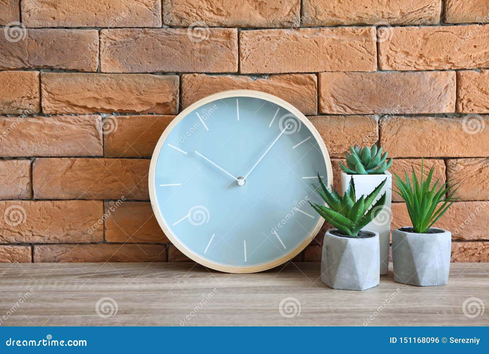 Modern Clock with Plants on Table Against Brick Wall Stock Photo