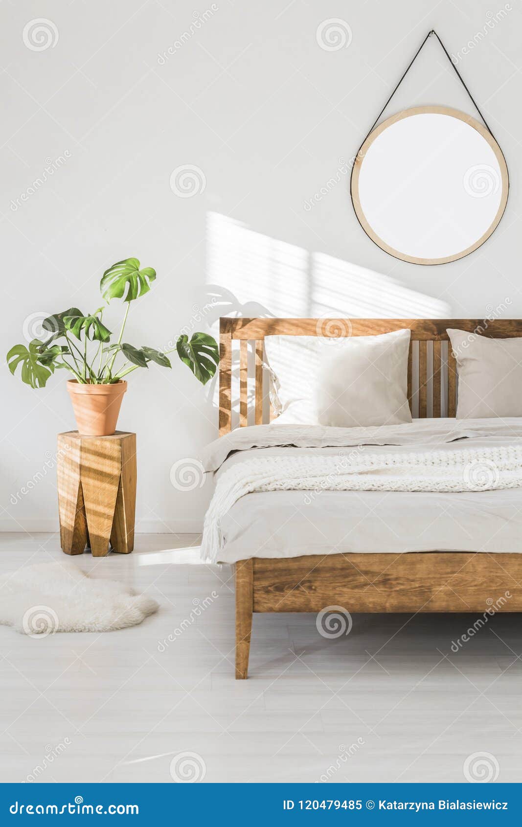 monstera plant on a tree trunk night stand and a round mirror on a white wall in a sunlit bedroom interior with wooden furniture