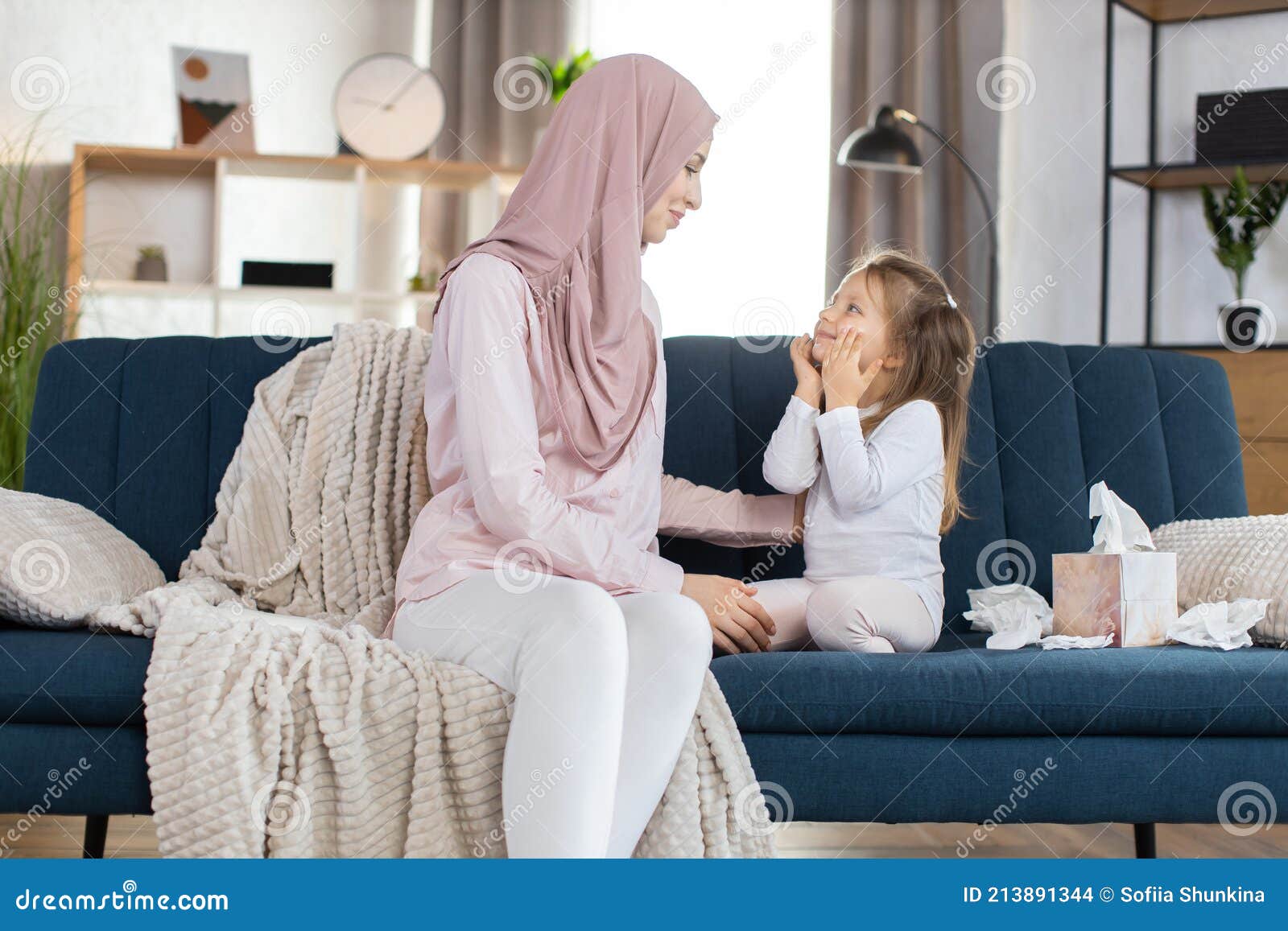 morning higiene routine at home. happy muslim mom in hijab, sits on sofa at cozy room and looks at her cute little