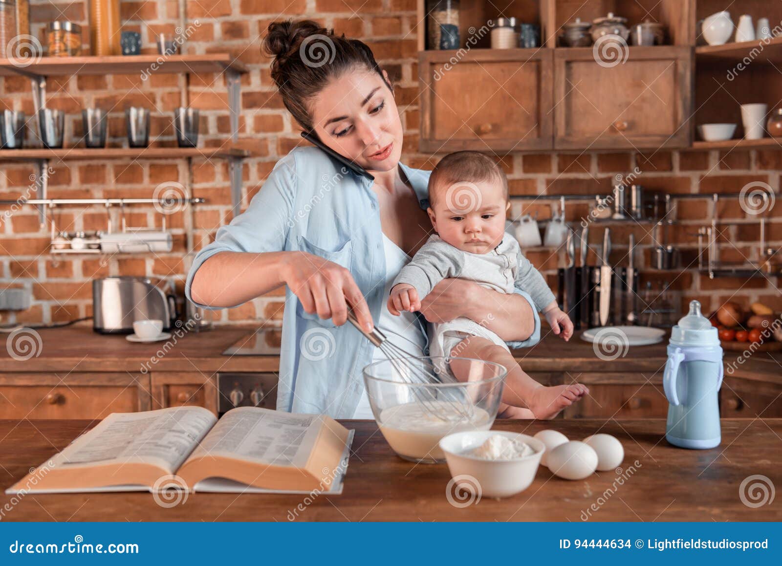 mother holding her son, talking on smartphone and mixing a dough at the kitchen. family life and multitasking concept