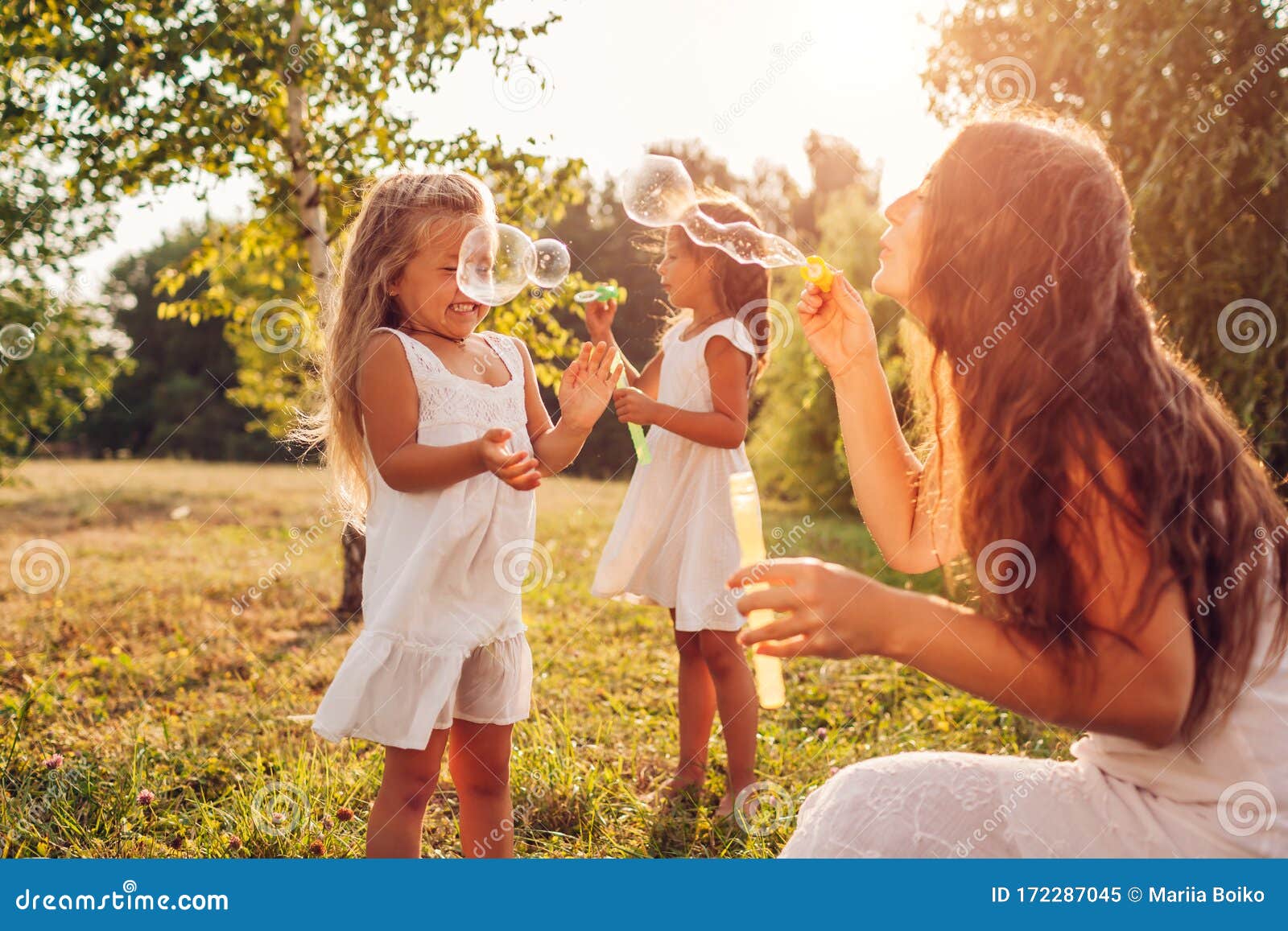 mother`s day. woman helps daughters to blow soap bubbles in summer park. kids having fun playing outdoors