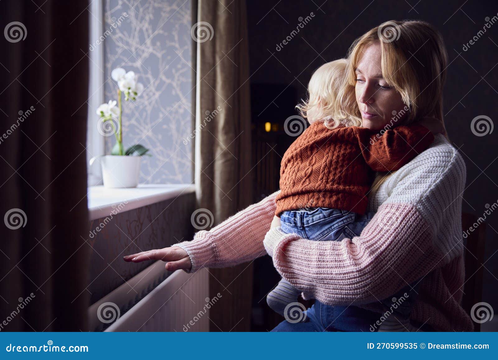 mother with son trying to keep warm by radiator at home during cost of living energy crisis