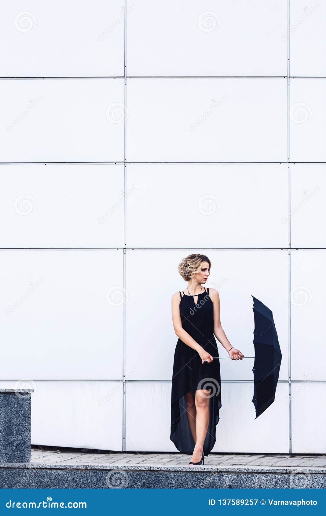 Moça elegante em um vestido preto e com um guarda-chuva que levanta contra uma parede cinzenta