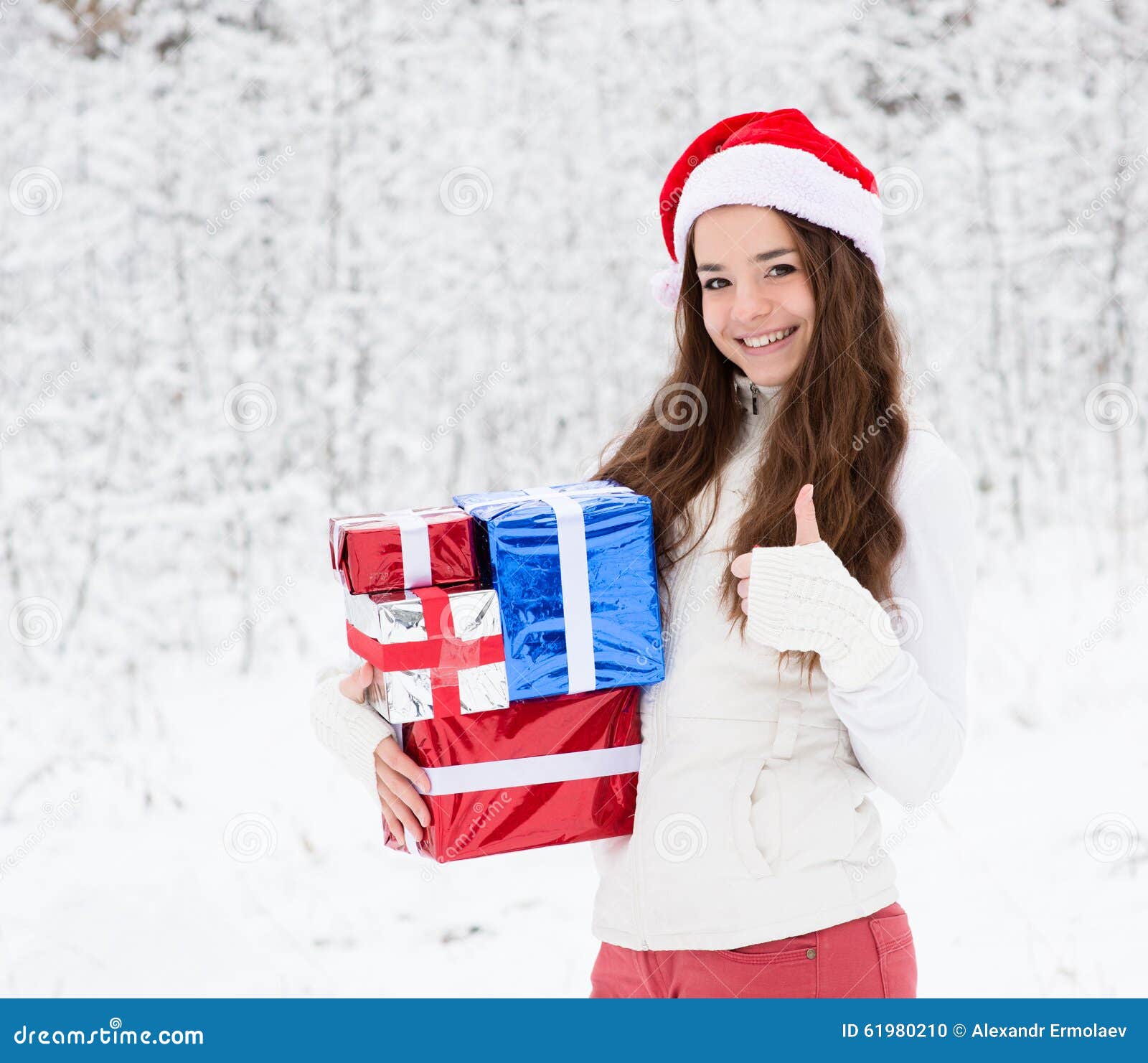 Muchacha adolescente con el sombrero de santa y las cajas de regalo rojas que muestran los pulgares para arriba en bosque del invierno