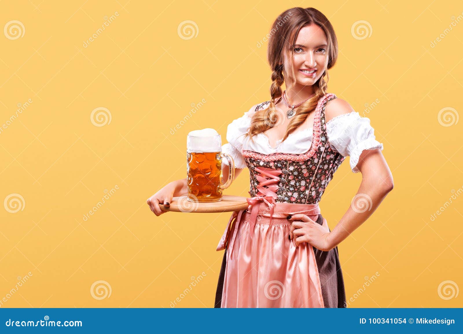 Muchacha atractiva joven de Oktoberfest - camarera, llevando un vestido bávaro tradicional, tazas de cerveza grandes de servicio. Retrato de medio cuerpo del blonde atractivo joven con el dirndl del color del pecho que lleva grande con la blusa blanca que sostiene la taza de cerveza en fondo anaranjado