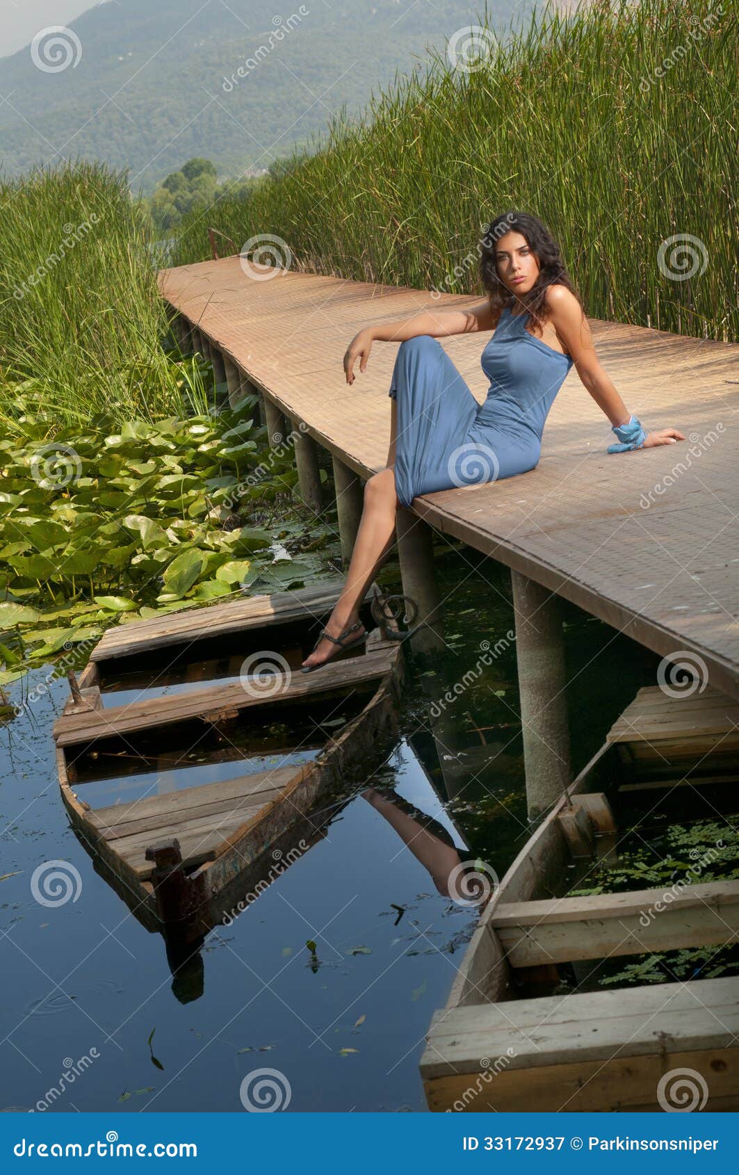 Muchacha atractiva que se sienta en un embarcadero. Foto vertical de una muchacha atractiva y hermosa, sentándose cerca de un barco exhausto en un lago. Ella se está sentando en un embarcadero. Los lirios y las cañas son visibles en el fondo.