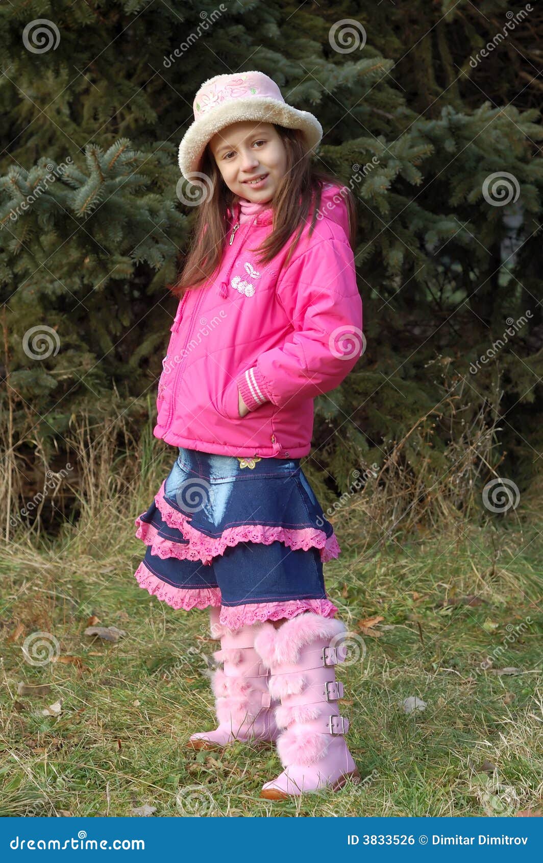 Muchacha feliz en campo. Chica joven feliz en capa, sombrero y cargador del programa inicial rosados en campo; árboles de pino en fondo.