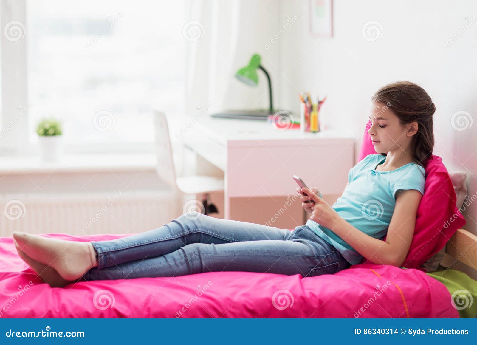 Muchacha sonriente que manda un SMS en smartphone en casa. Concepto de los niños, de la tecnología y de la comunicación - muchacha sonriente que manda un SMS en smartphone y que miente en cama en casa