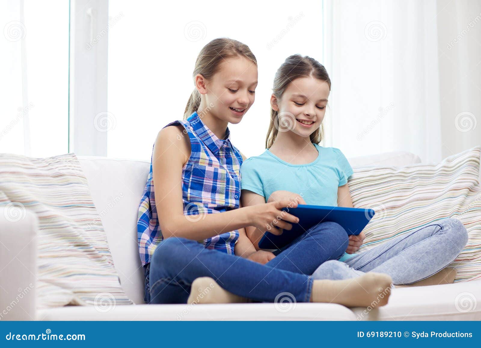 Muchachas felices con la PC de la tableta que se sienta en el sofá en casa. Gente, niños, tecnología, amigos y concepto de la amistad - niñas felices con el ordenador de la PC de la tableta que se sienta en el sofá en casa