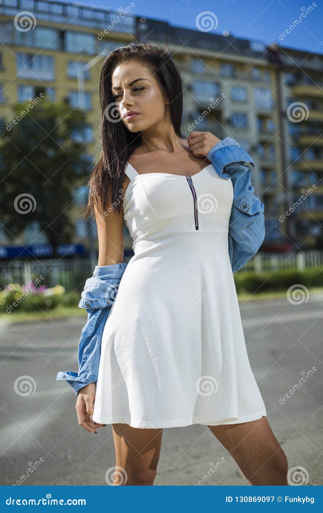 Mujer Bonita Atractiva En El Vestido Blanco Con La Chaqueta De Los Vaqueros Que Mira La Cámara Mientras Que Se Coloca de archivo Imagen de calle, mirando: 130869097