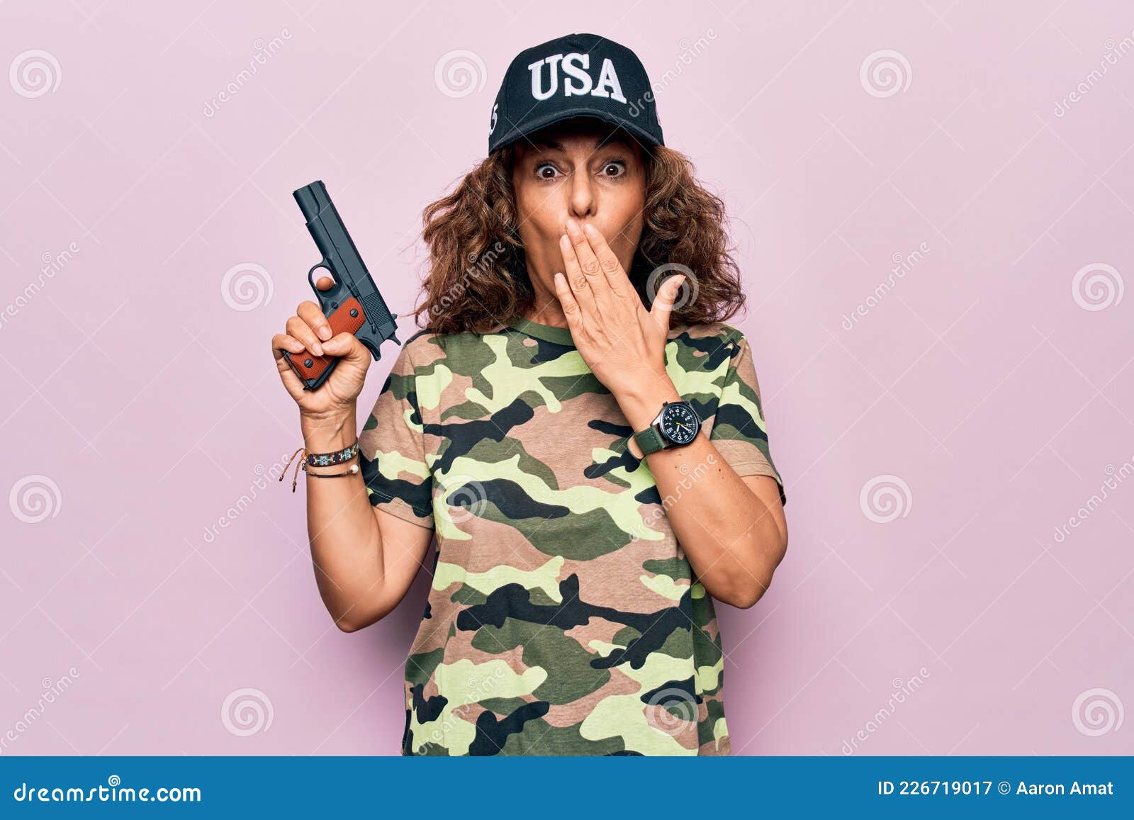 Mujer De Mediana Edad Guapa Cazadora Con Camiseta De Camuflaje Y Gorra De Ee.uu. Sosteniendo Una Pistola Que Cubría La Boca Con La Imagen de archivo cazador, 226719017