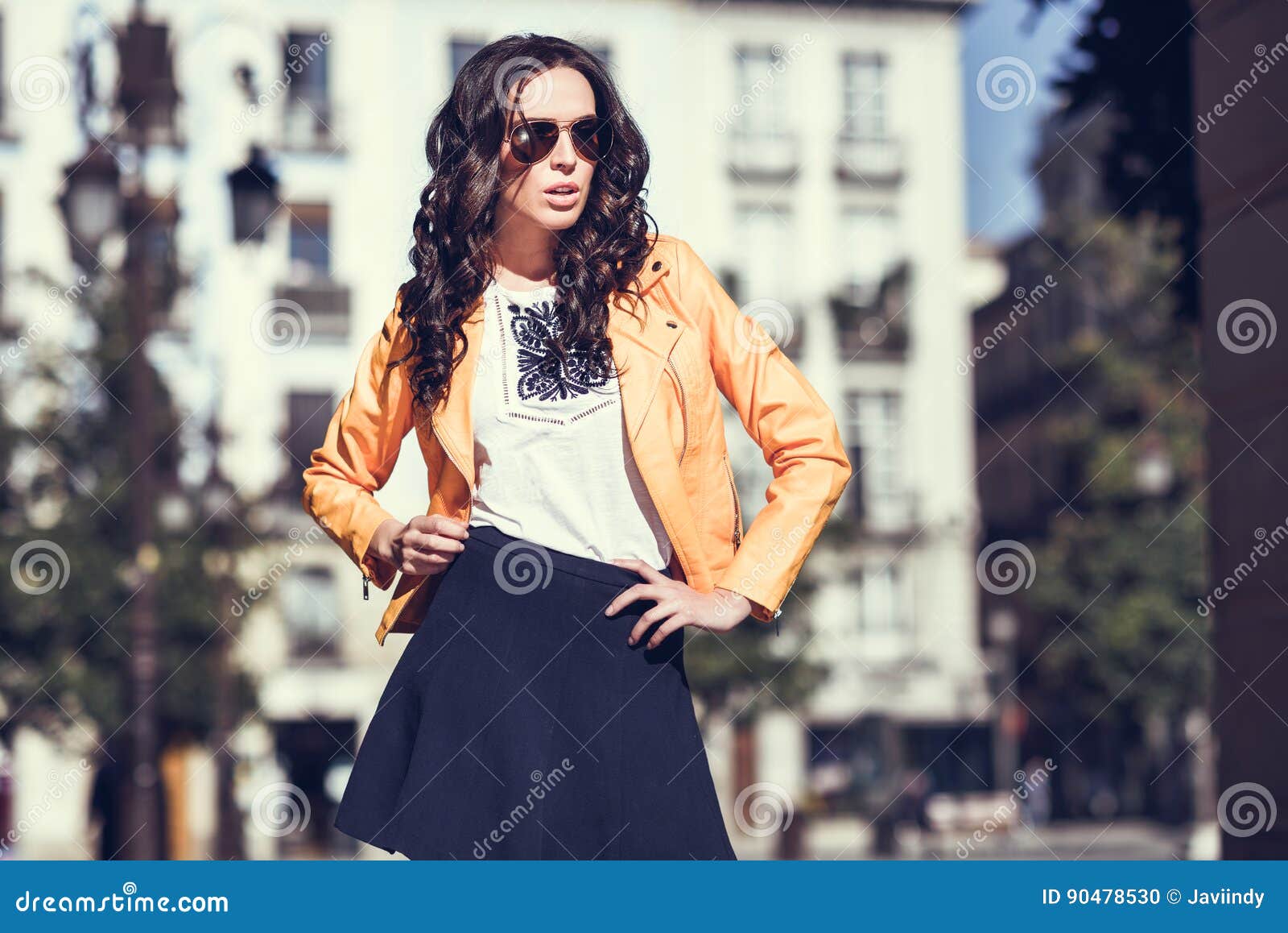 Mujer morena joven con las gafas de sol en fondo urbano. Mujer morena joven con las gafas de sol Muchacha, modelo de la moda, chaqueta moderna anaranjada que lleva y falda azul, colocándose en fondo urbano