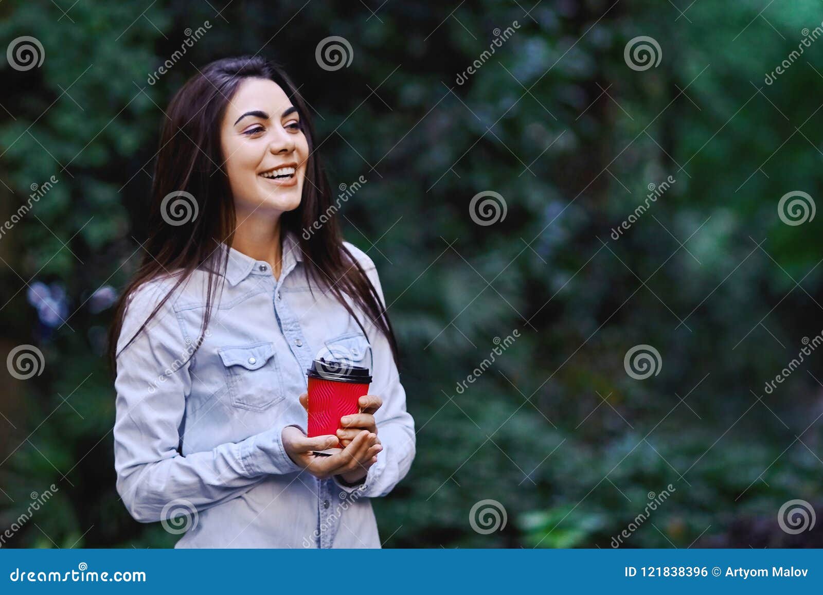 Mujer sonriente joven que sostiene la taza de café. Mujer morena sonriente joven, camisa azul del dril de algodón que lleva y sostener la taza de café roja