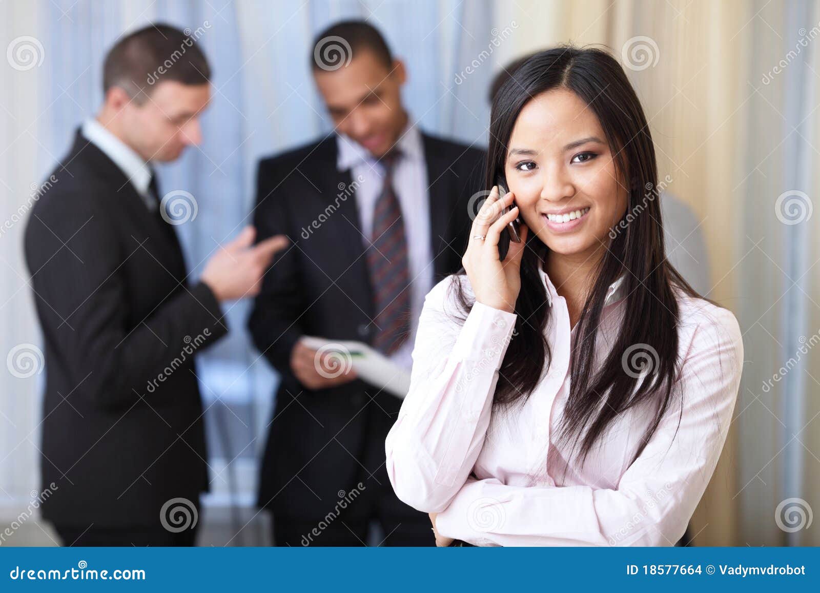 Mulher de negócios asiática nova no telefone. Retrato da mulher asiática feliz nova no telefone com seus sócios comerciais no fundo