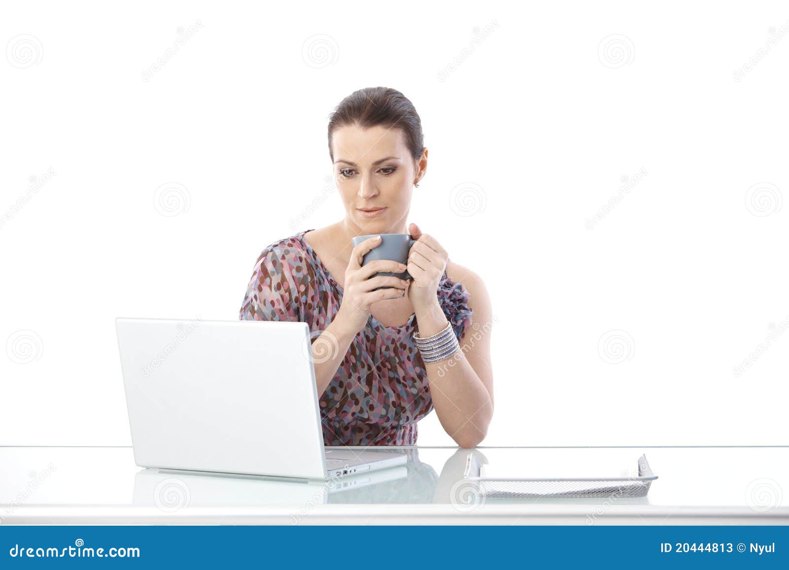 Mulher do Meados de-adulto que usa o portátil. Mulher do Meados de-adulto que senta-se na mesa que olha a tela do computador portátil, prendendo a caneca de café, fundo branco.