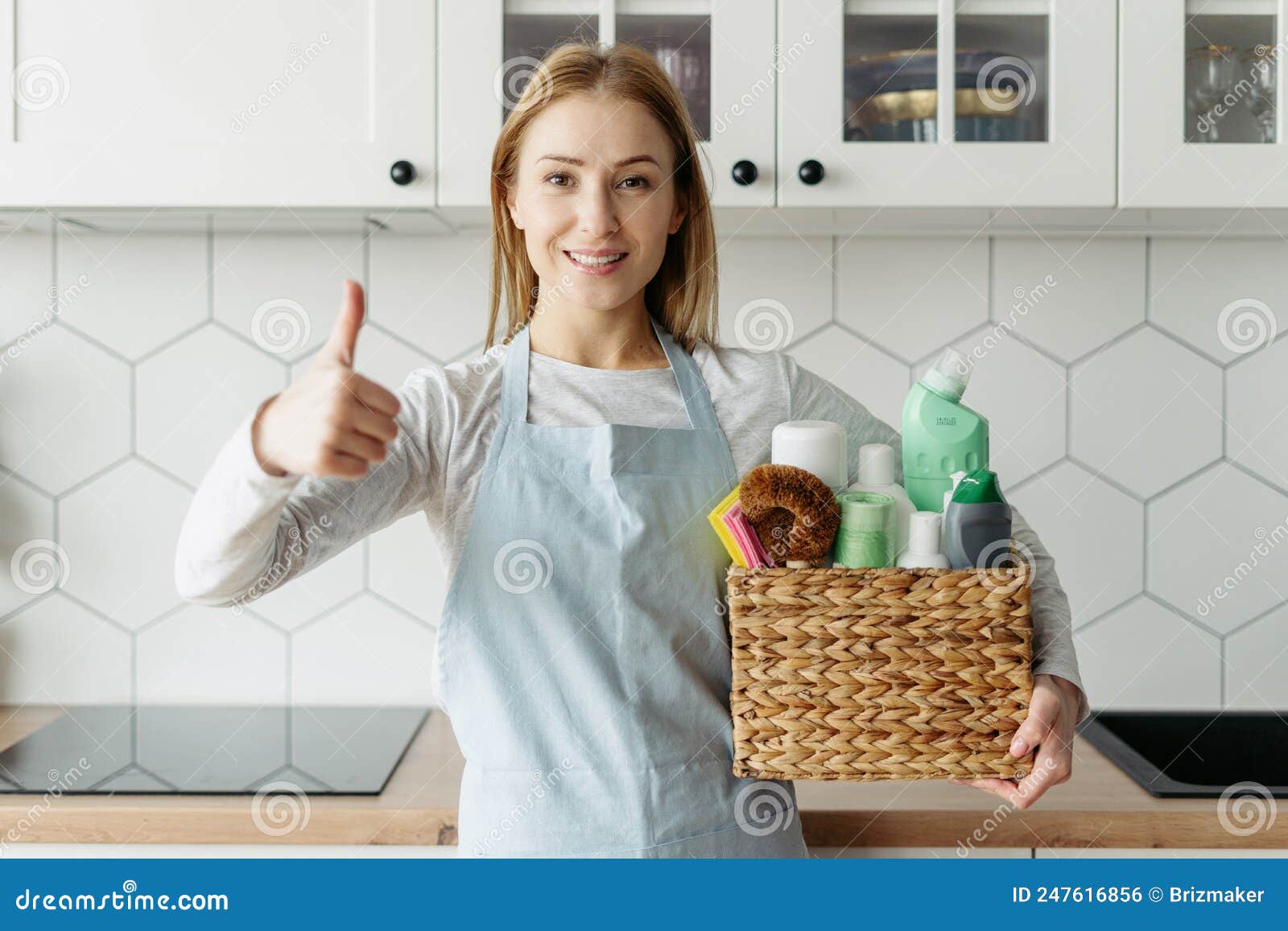Mulher recomenda produtos de limpeza mostrando polegares para cima. Linda mulher em avental segurando uma caixa com detergentes e trapos prontos para limpar sua casa. trabalhador de serviços de limpeza recomenda produtos químicos domésticos mostrando polegares para cima e sorrindo publicidade ampla