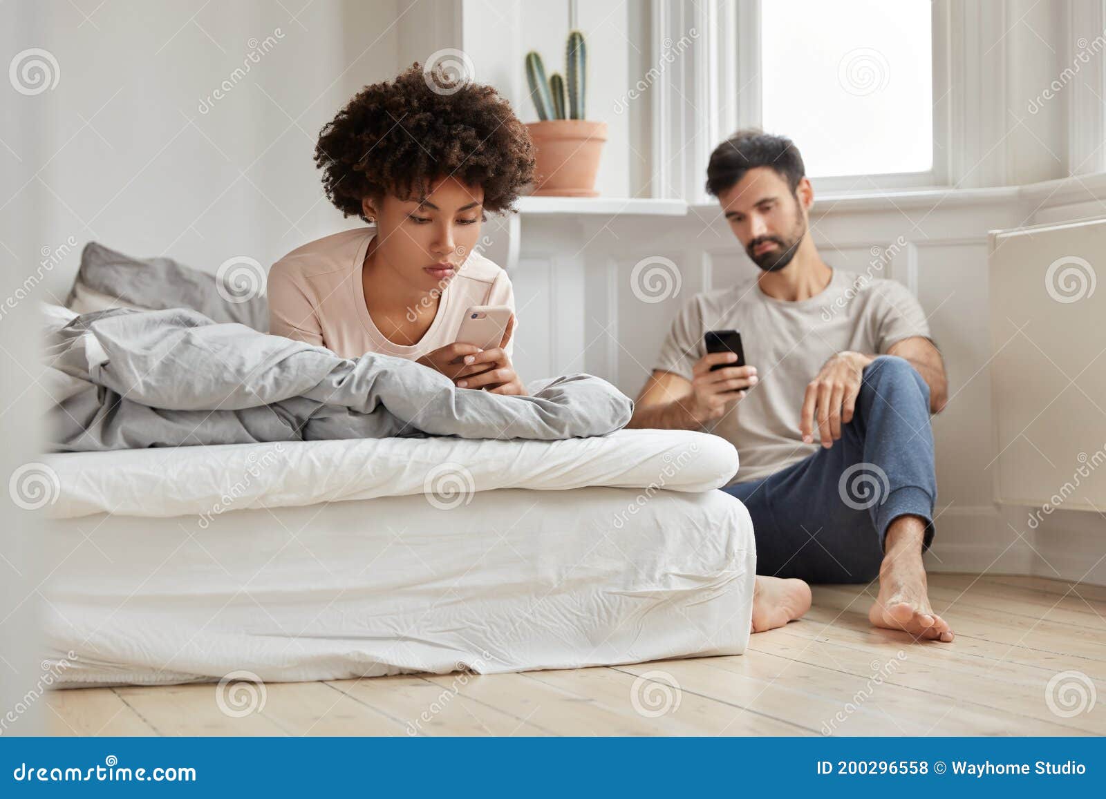 multiethnic couple watch video via social networks, hold modern cellulars, wife lies in bed, husband sits near at floor