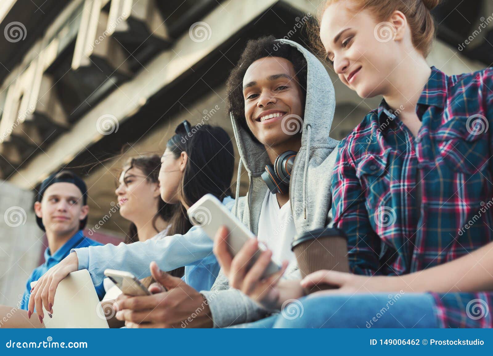 multiethnic teen friends sitting outdoors, talking and using gadgets