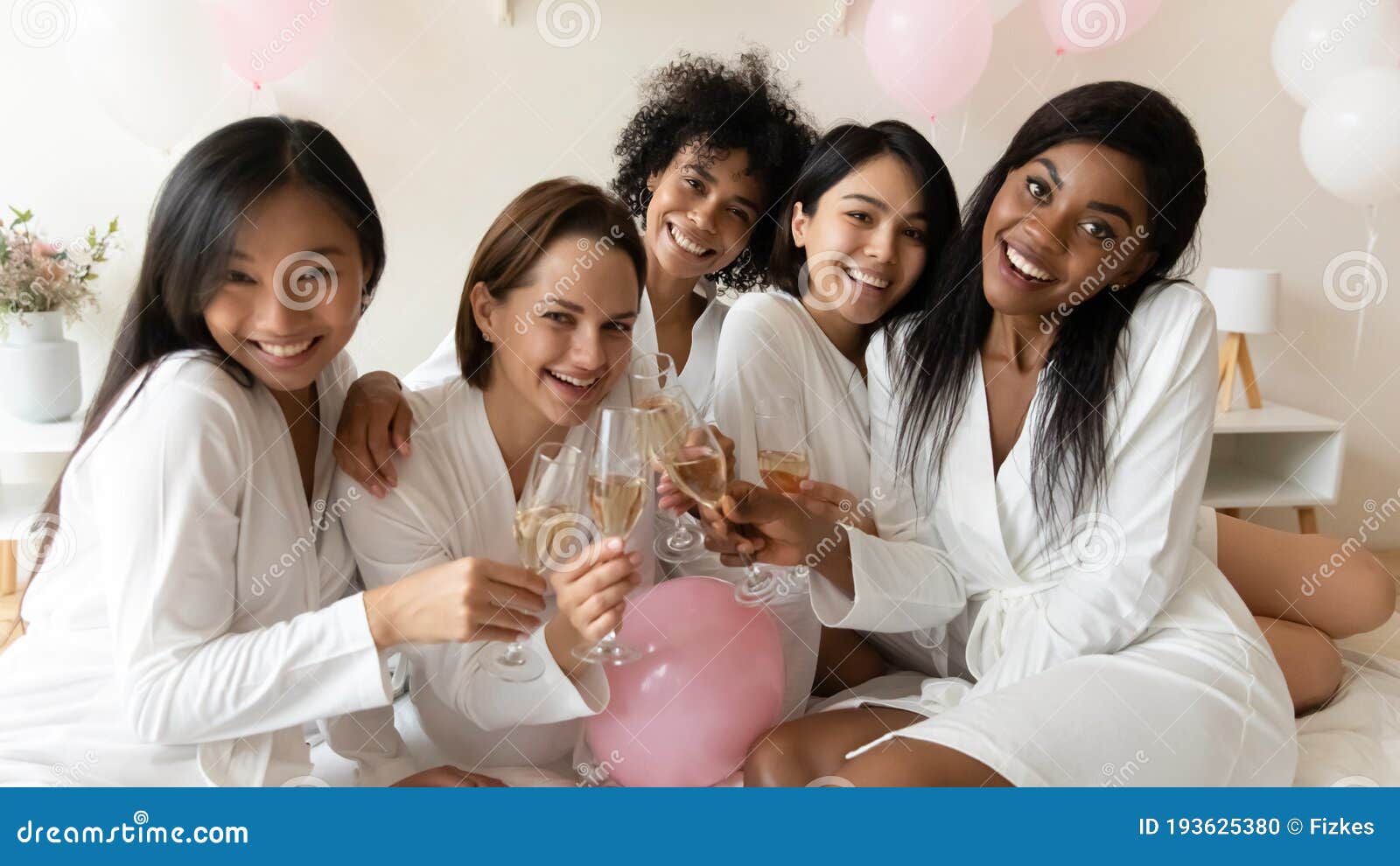 multiracial girls in bathrobes holding champagne glasses celebrating bachelorette party