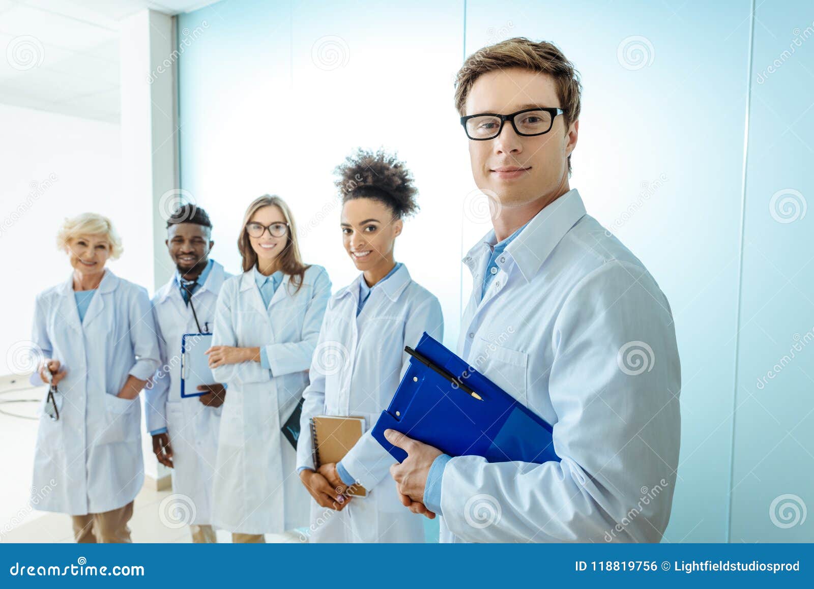 multiracial group of smiling medical interns in lab coats standing in a row with clipboards