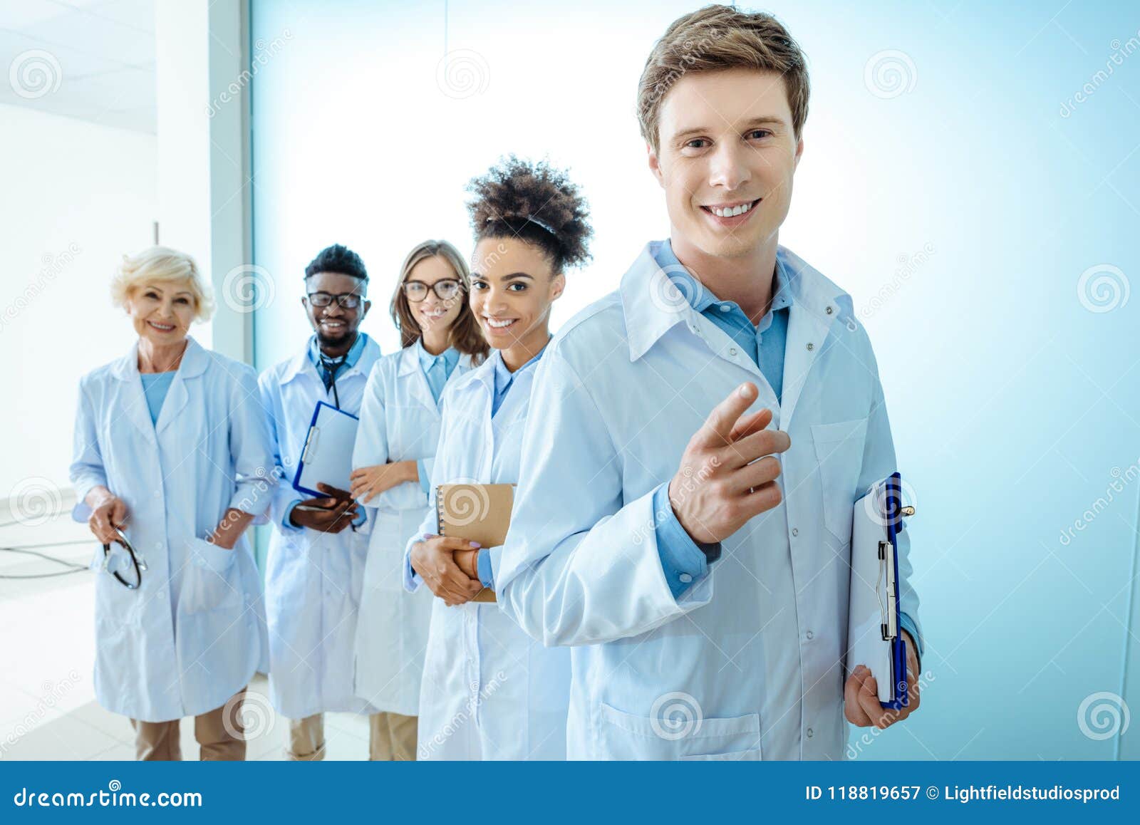 multiracial group of smiling medical interns in lab coats standing in a row with clipboards