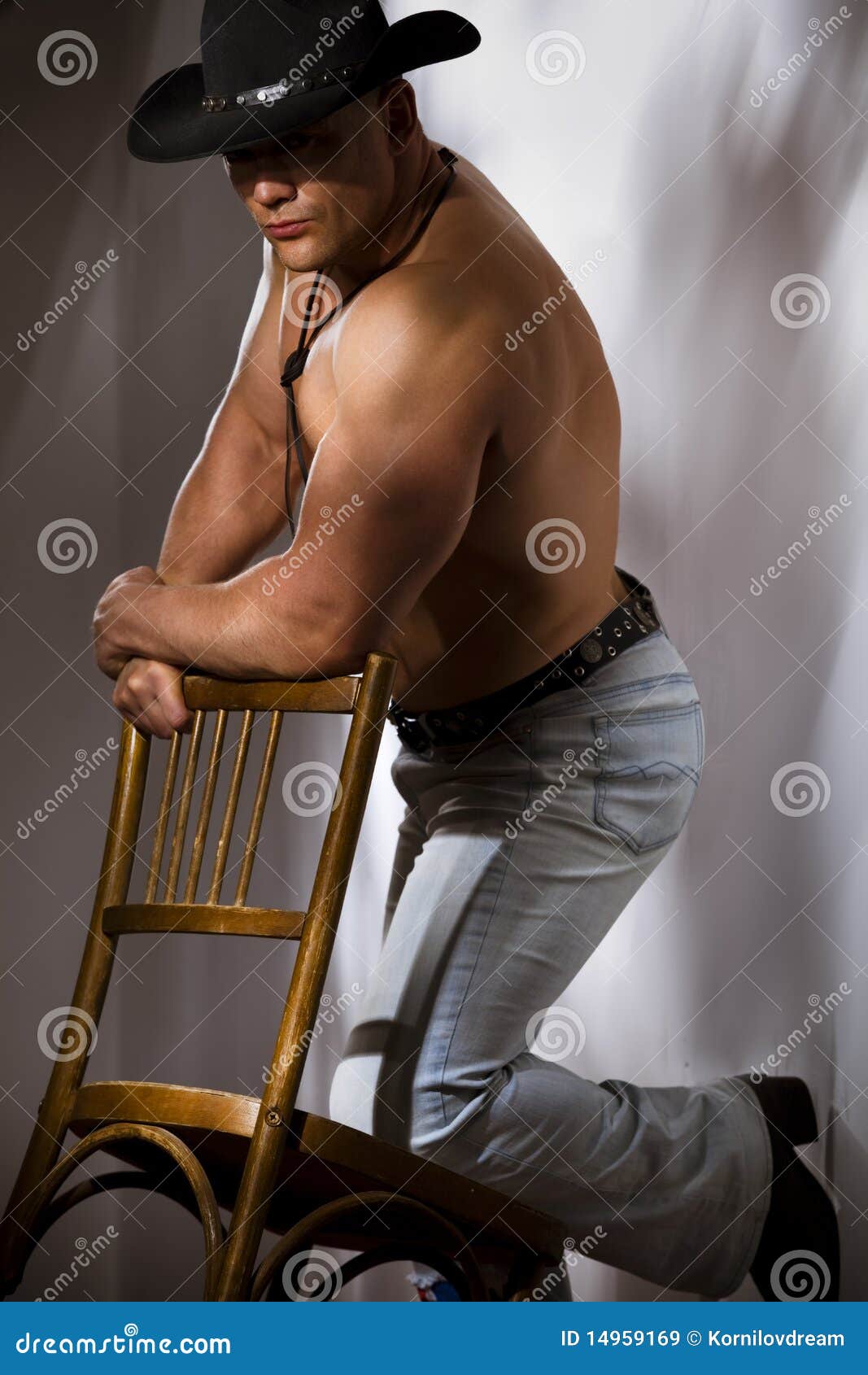 Photo about Muscular shirtless cowboy leaning on chair against a white wall. 