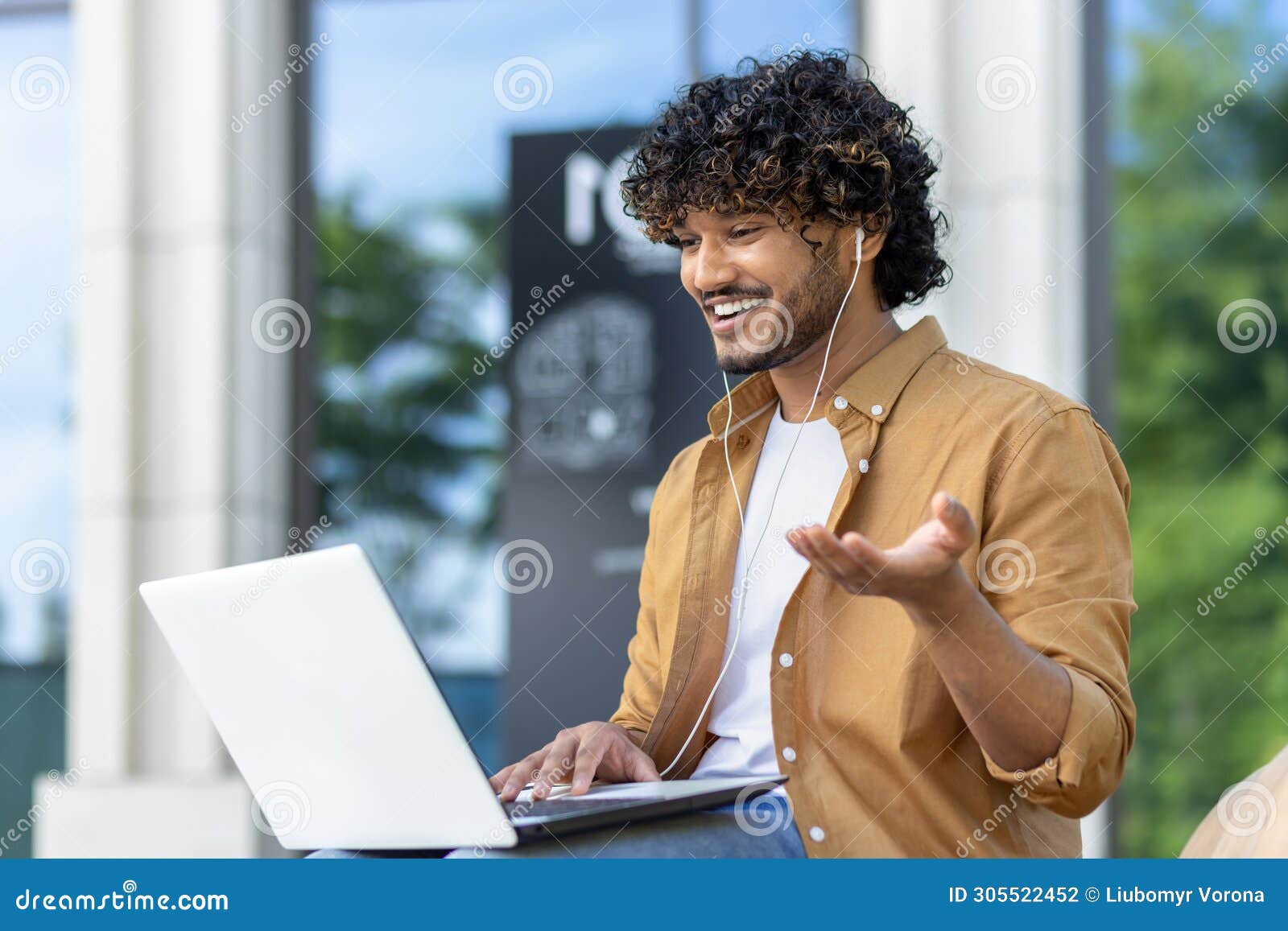 muslim young man sitting on a bench on a city street wearing headphones and smilingly talking on a video call on a