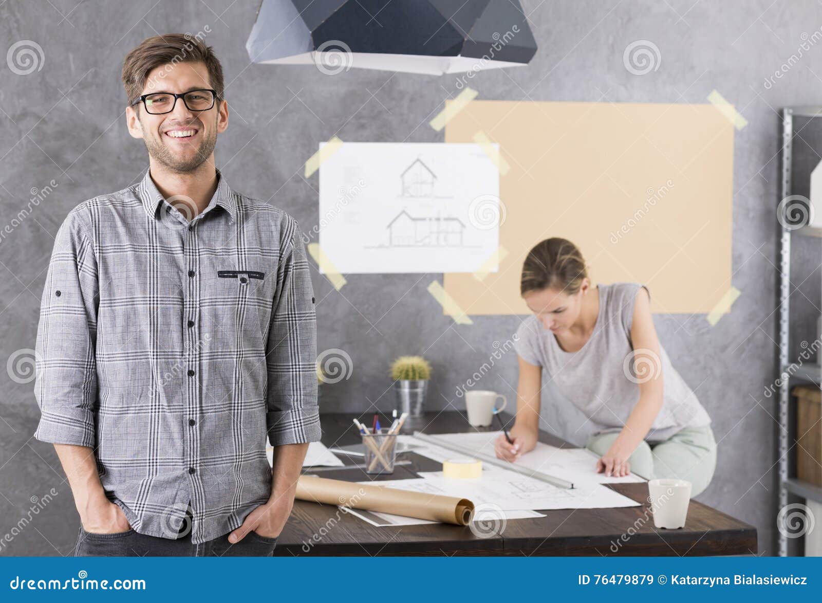 My work makes me happy. Close shot of young smiled men at the office with the women sitting at a desk and working on project behind him