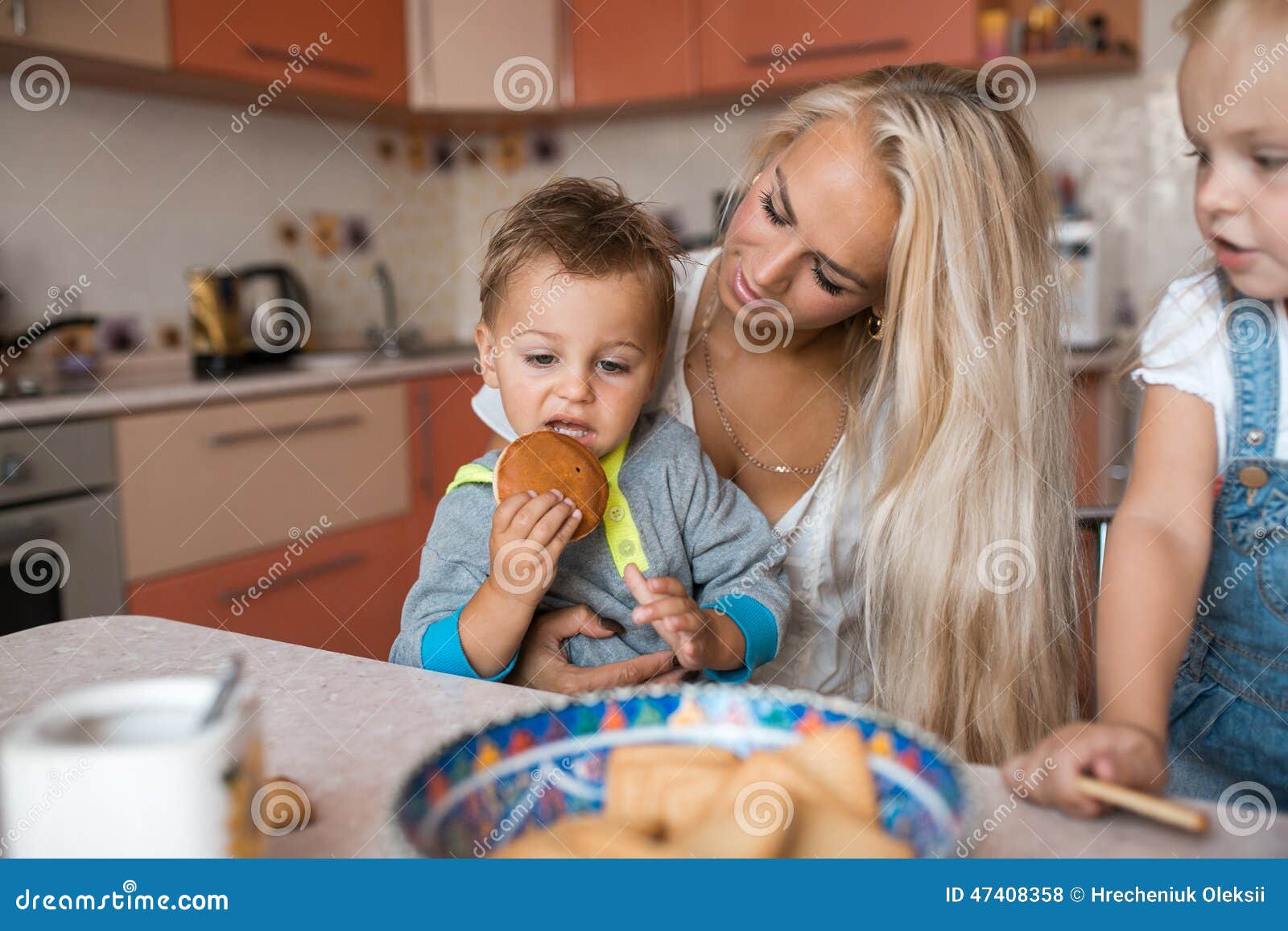 A mãe com as crianças na cozinha, filho come uma cookie. A mãe com a família que tem o divertimento na cozinha, filho come uma cookie