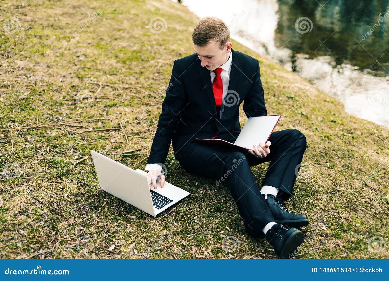 M?ody ucze? studiuje na naturze w parku blisko jeziora facet w czarnym kostiumu czerwonym krawacie i czyta laptop i pisze w notatniku