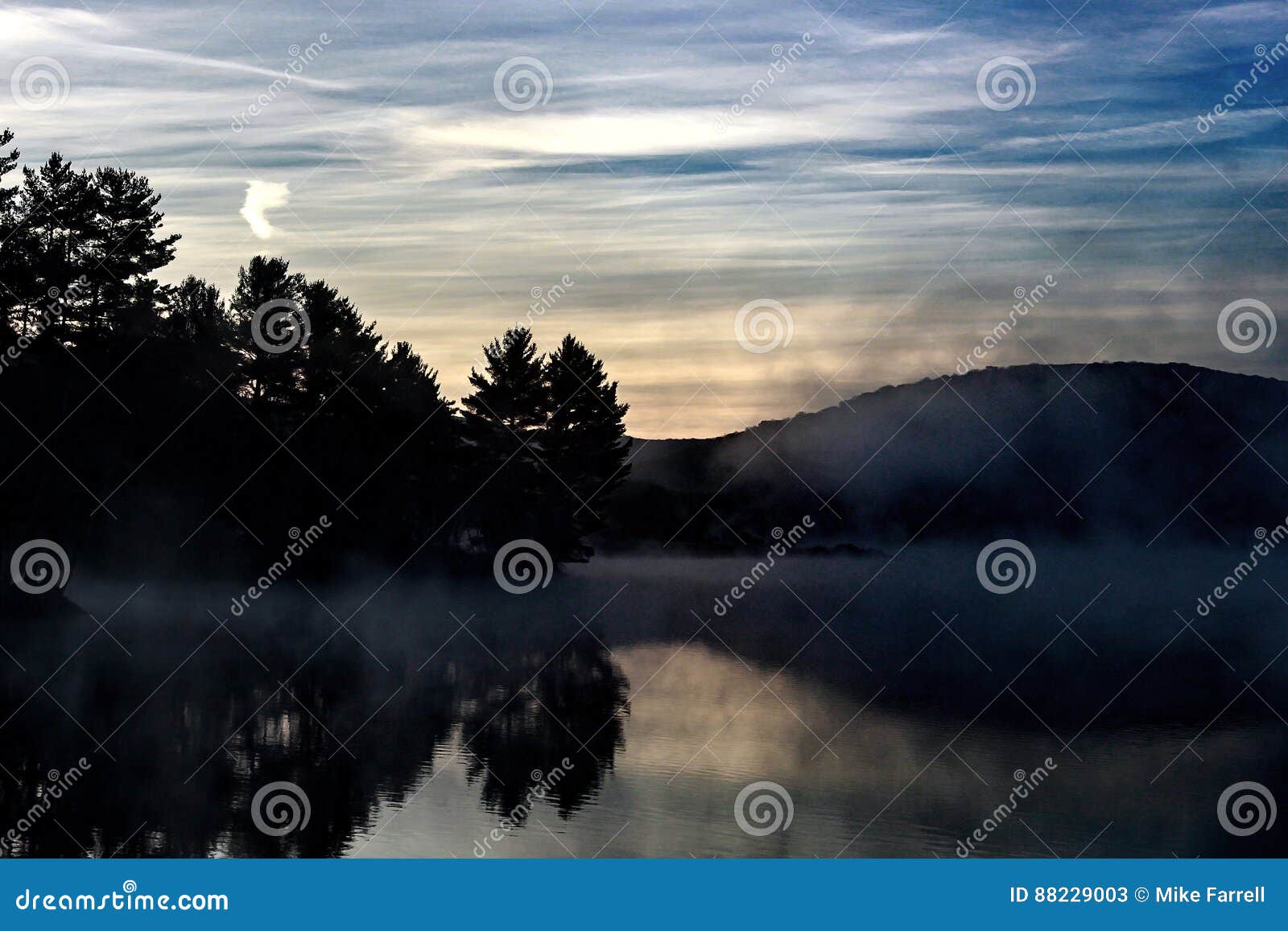 Nascer do sol nevoento sobre um lago com algumas árvores e montanhas