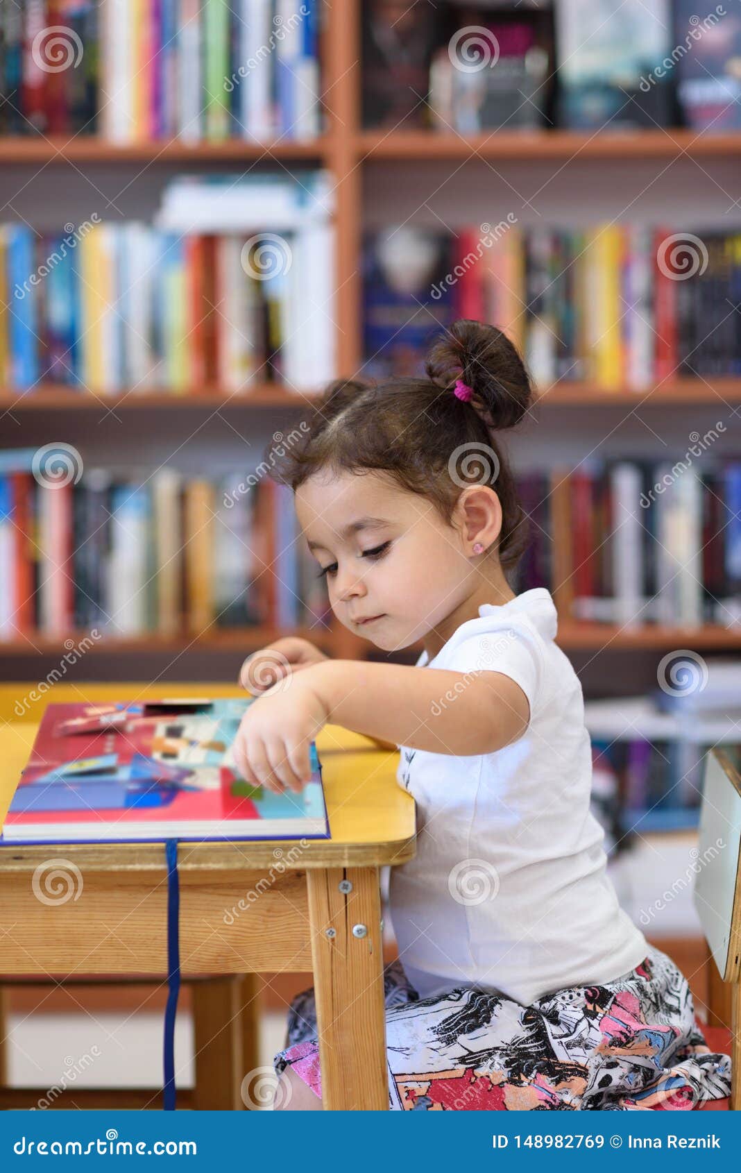 Ni?a feliz del ni?o que lee un libro. Little Girl Indoors In Front Of Books. Cute Young Toddler Sitting On A Chair Near Table and Reading Book. Child reads in a bookstore, surrounded by colorful books. Library, Shop, Shelving In Home.