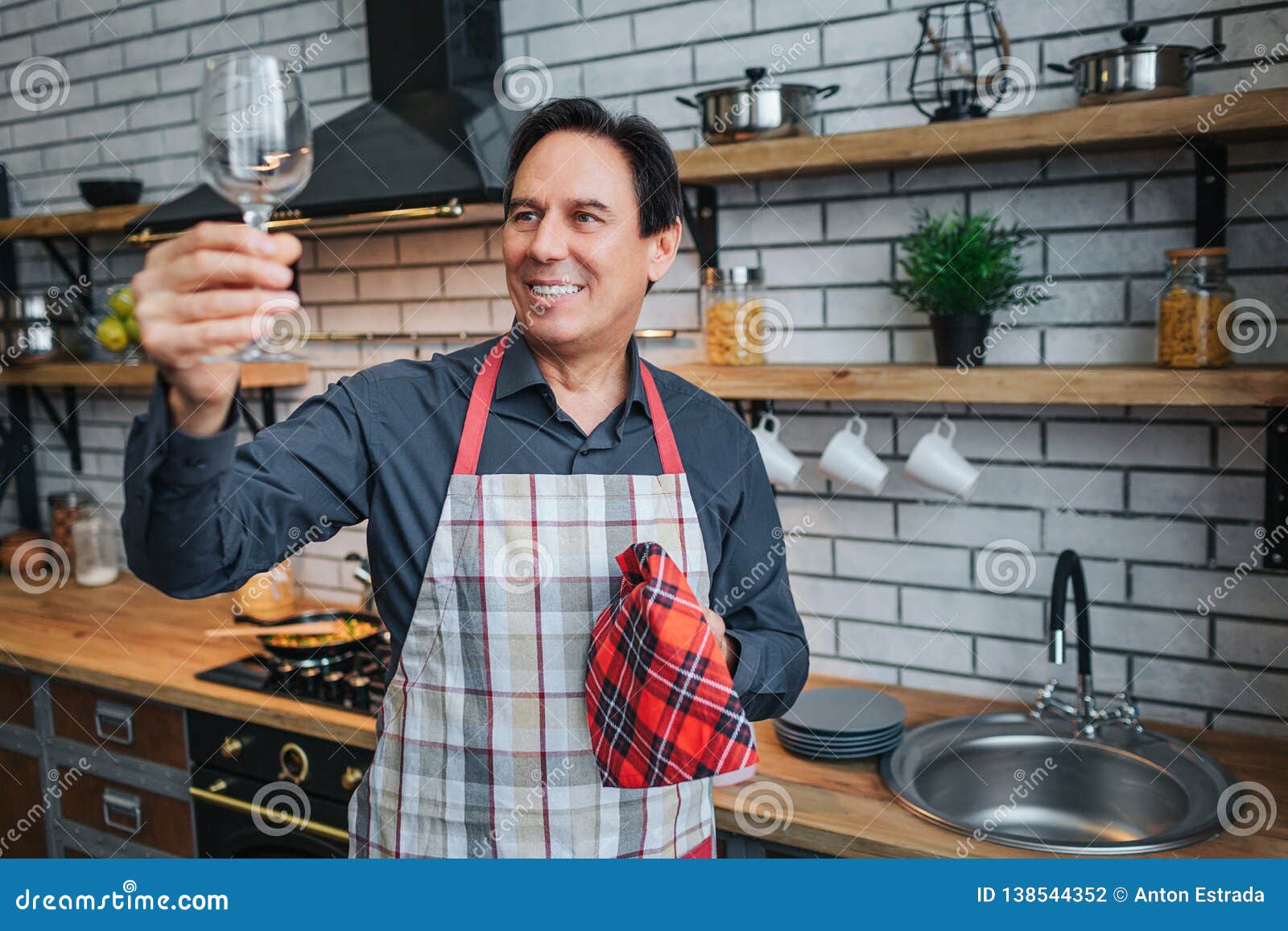 Nice Adult Man In Apron Stand Alone And Look At Glass In Kitchen