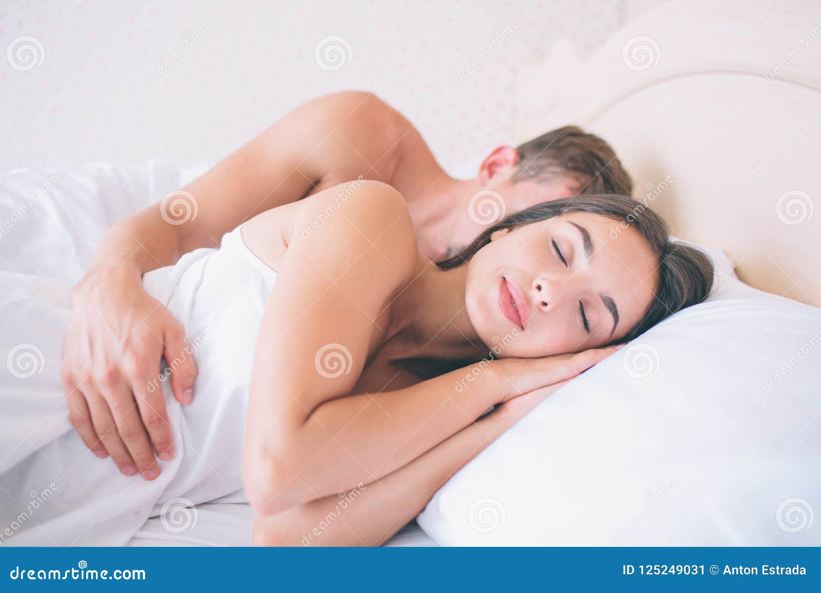 nice and beautiful girl sleeps in fresh and white bed. she keeps hands under head. guy is lying behinds her. he sleeps