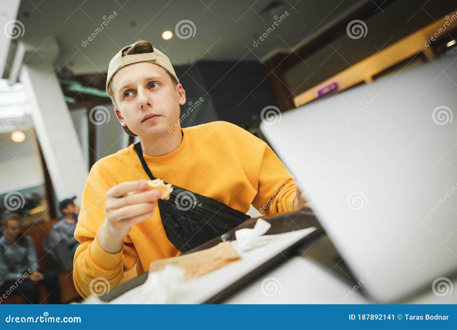 nice guy is working on a laptop and eats in a fast food restaurant, looking sideways with a serious face. young man sitting in a