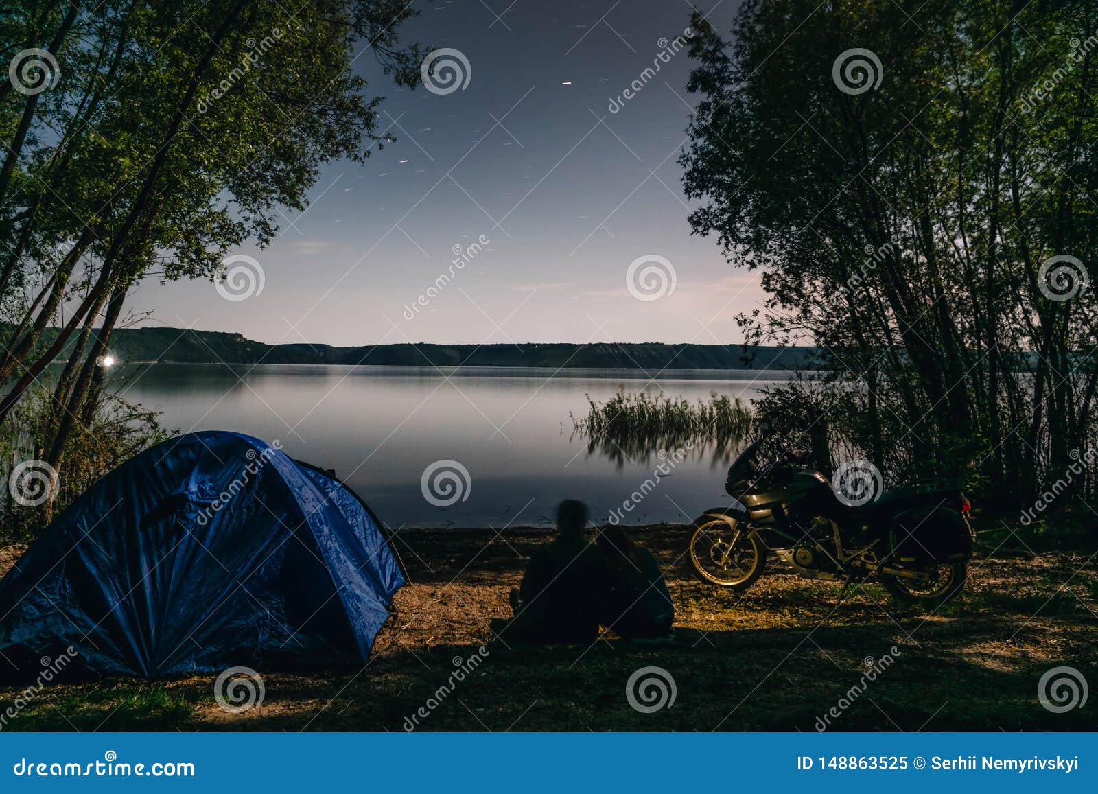 night camping on lake shore. man and woman is sitting. couple tourists enjoying amazing view of night sky full of stars. blue tent