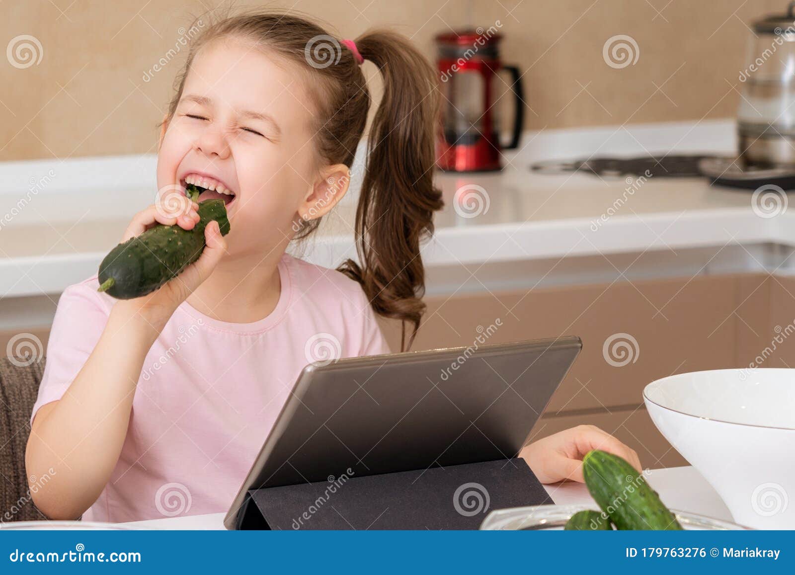 Niña Pequeña Comiendo Pepino Y Usando El Tablet En La Cocina Foto De