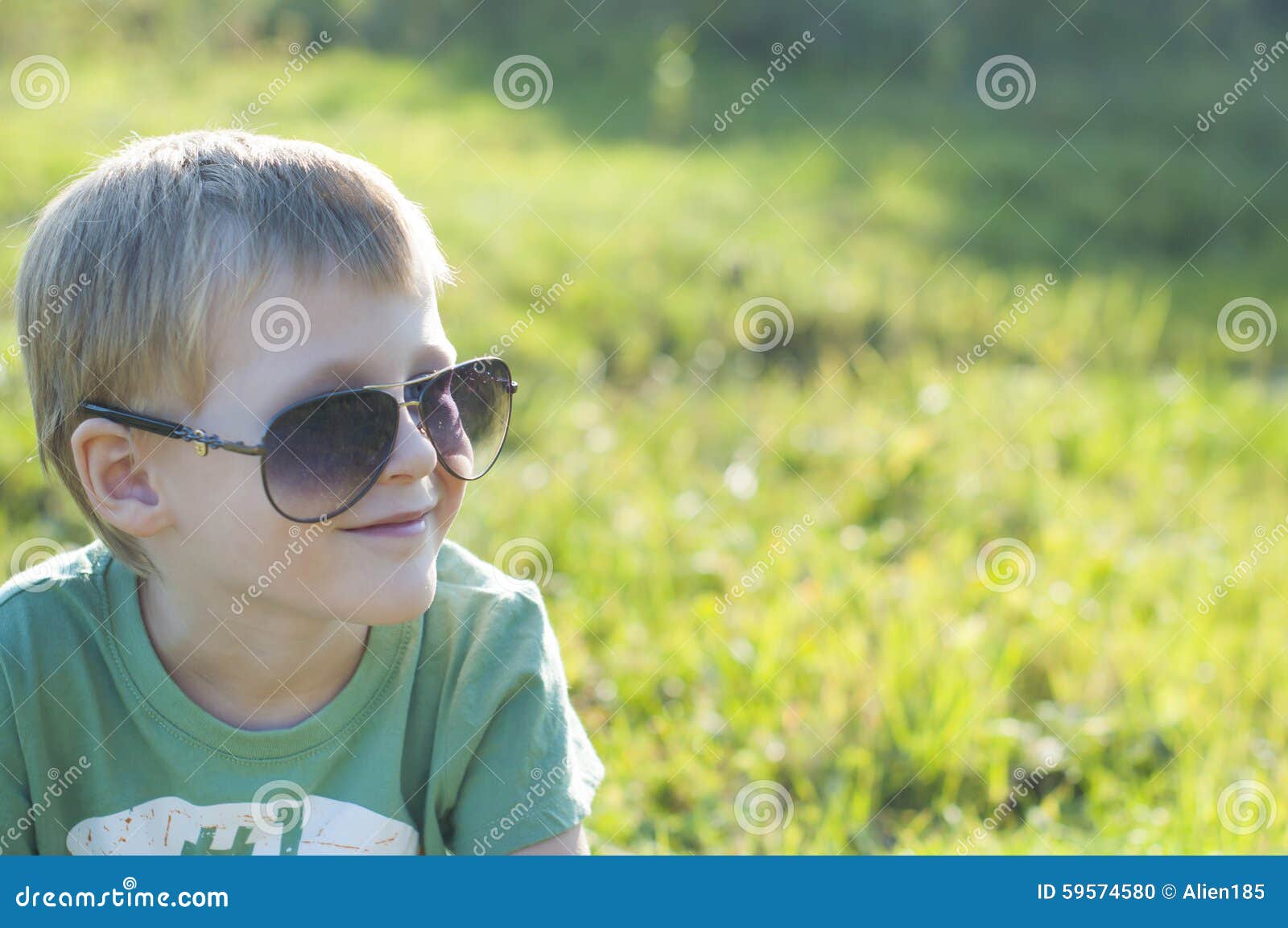 Niño adorable lindo en las gafas de sol que se sientan en hierba verde en el parque en un día maravilloso soleado