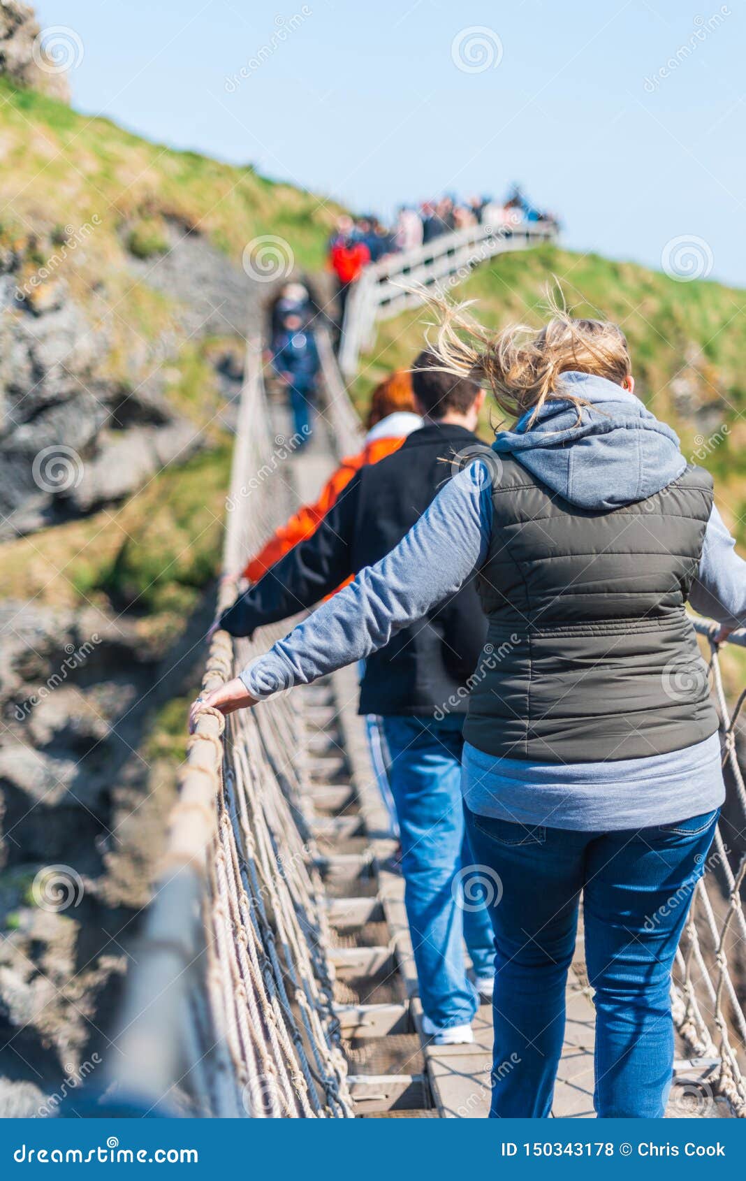 NORDIRLAND, GROSSBRITANNIEN - 8. APRIL 2019: Erschrockene Touristen kreuzen die gefährliche aber schöne Carrick-a-Redeseil-Brücke. Leute schauen unten und halten an, während sie die Seilbrücke in Irland an einem windigen Tag im April kreuzen