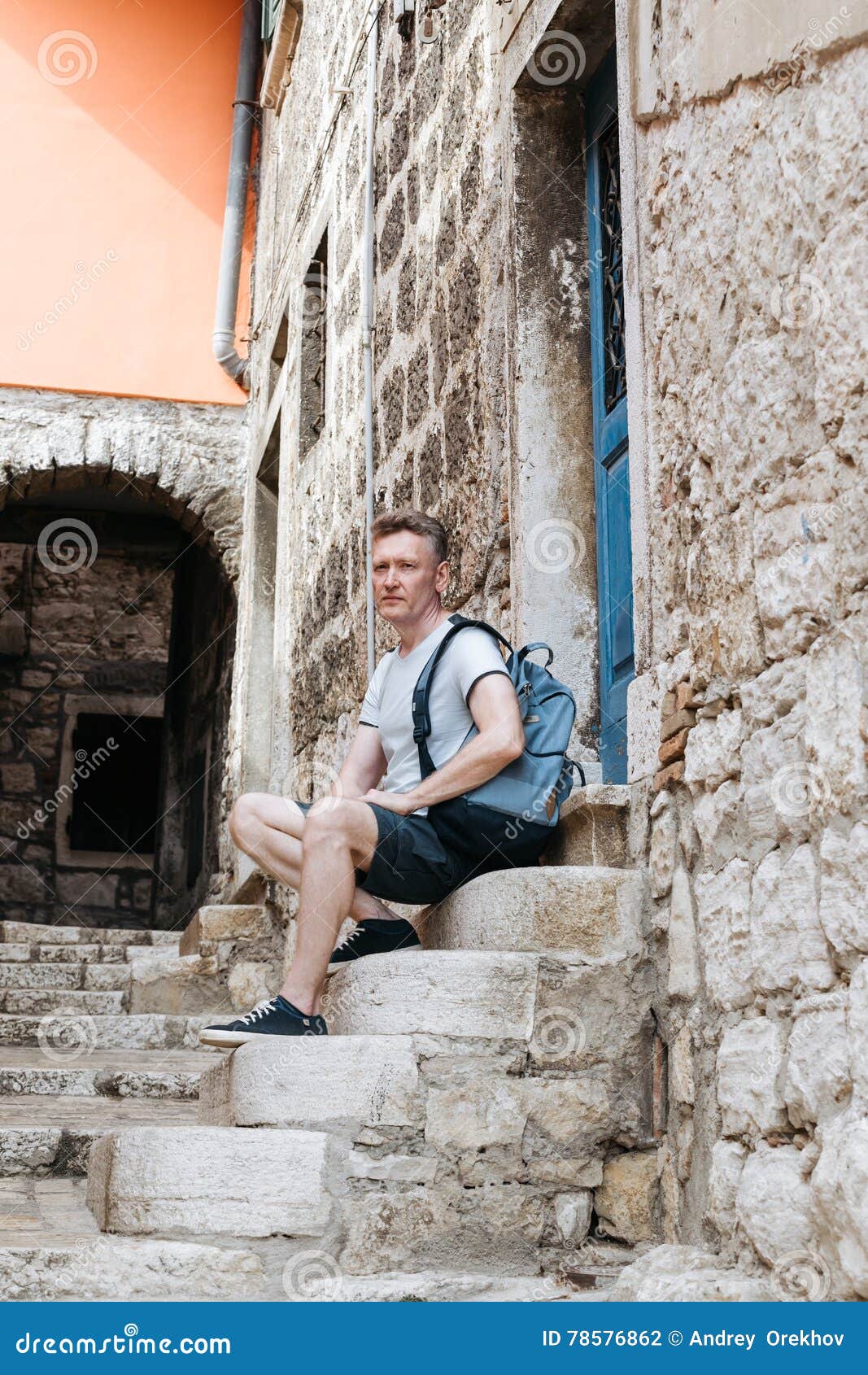 O homem à moda do turista vestiu-se em uma camisa branca e no short com a trouxa sobre seu ombro Assento nas etapas da cidade eur. O homem à moda do turista vestiu-se em uma camisa branca e em um short azul com uma trouxa azul sobre seu ombro Assento nas etapas de uma cidade europeia velha