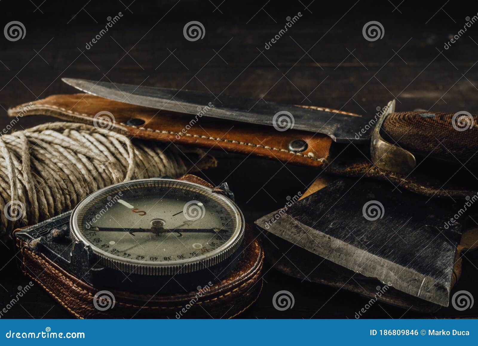 old military compass, rusty hunting bushcraft knife, small axe and a linen rope on the dark wooden table.