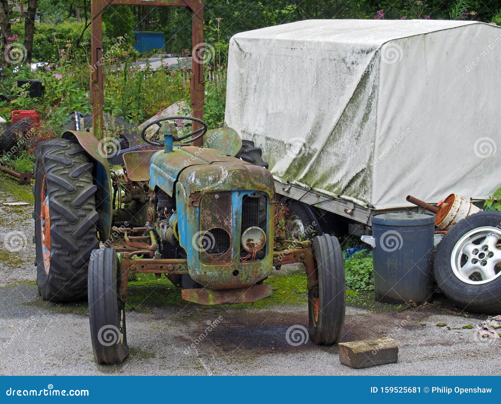 An old rusting abandoned tractor next to a dirty tarpaulin covered trailer and junk in a farmyard