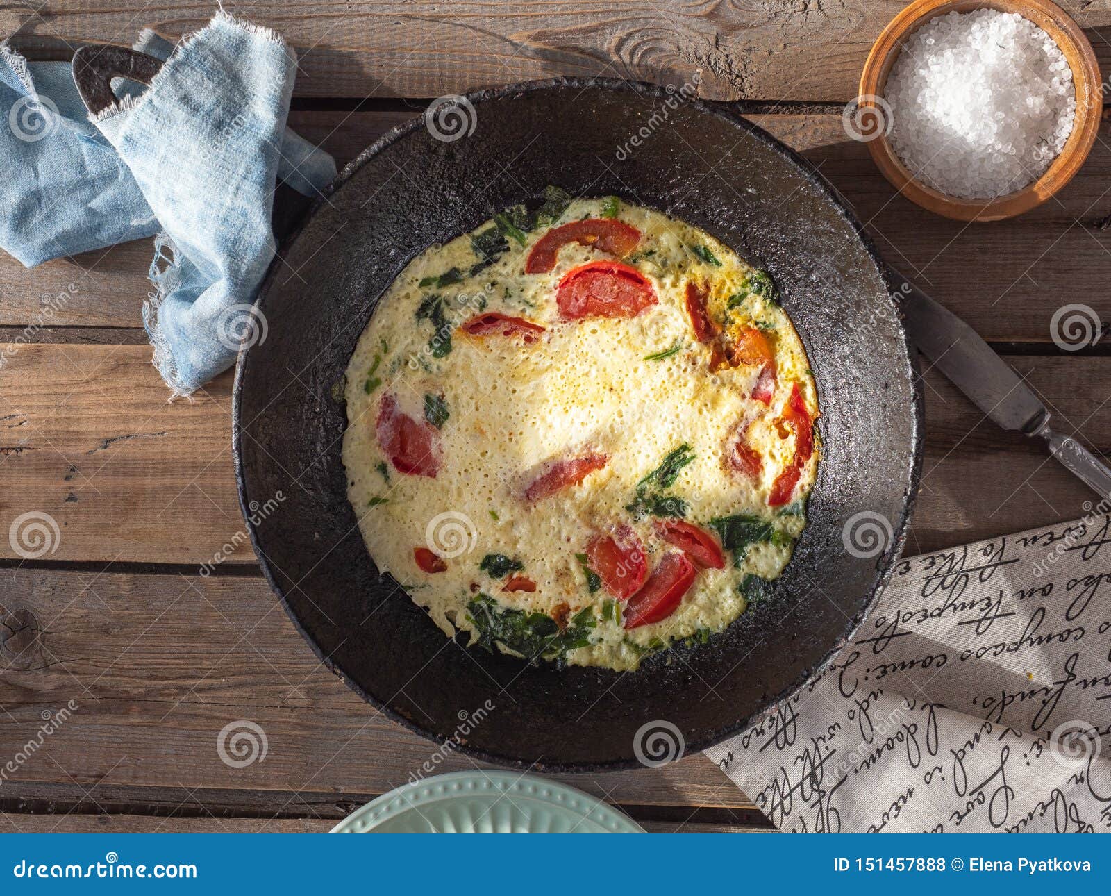 Omelette con i pomodori in una pentola rotonda del ghisa su una vecchia tavola del bordo, sui tovaglioli del cotone e sul sale grosso in un agitatore di sale dell'argilla, coltello da tavola