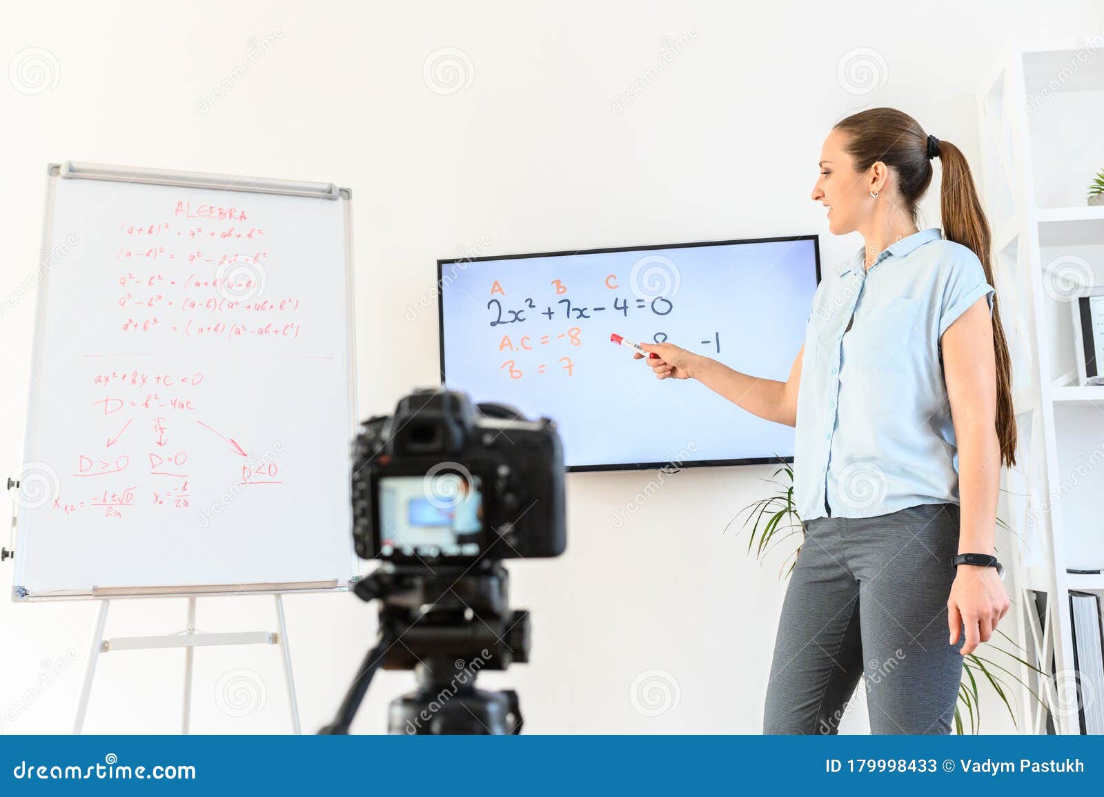 female teacher is recording classes on a camera, she stands near flip chart and screen with a marker in hand, camera in foreground
