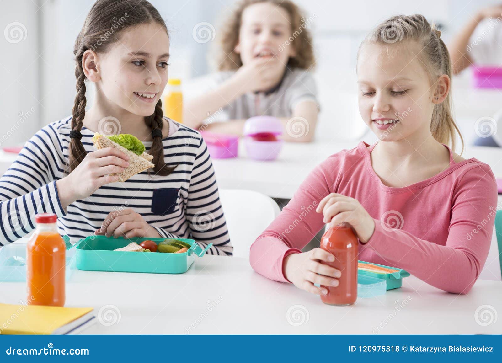 Opinião dianteira duas meninas júniors em um bar de escola durante o lunc. Opinião dianteira duas meninas júniors em um bar de escola durante a pausa para o almoço Um que guarda um sanduíche do wholewheat, o outro que abre uma garrafa do suco vegetal orgânico Outras crianças no fundo